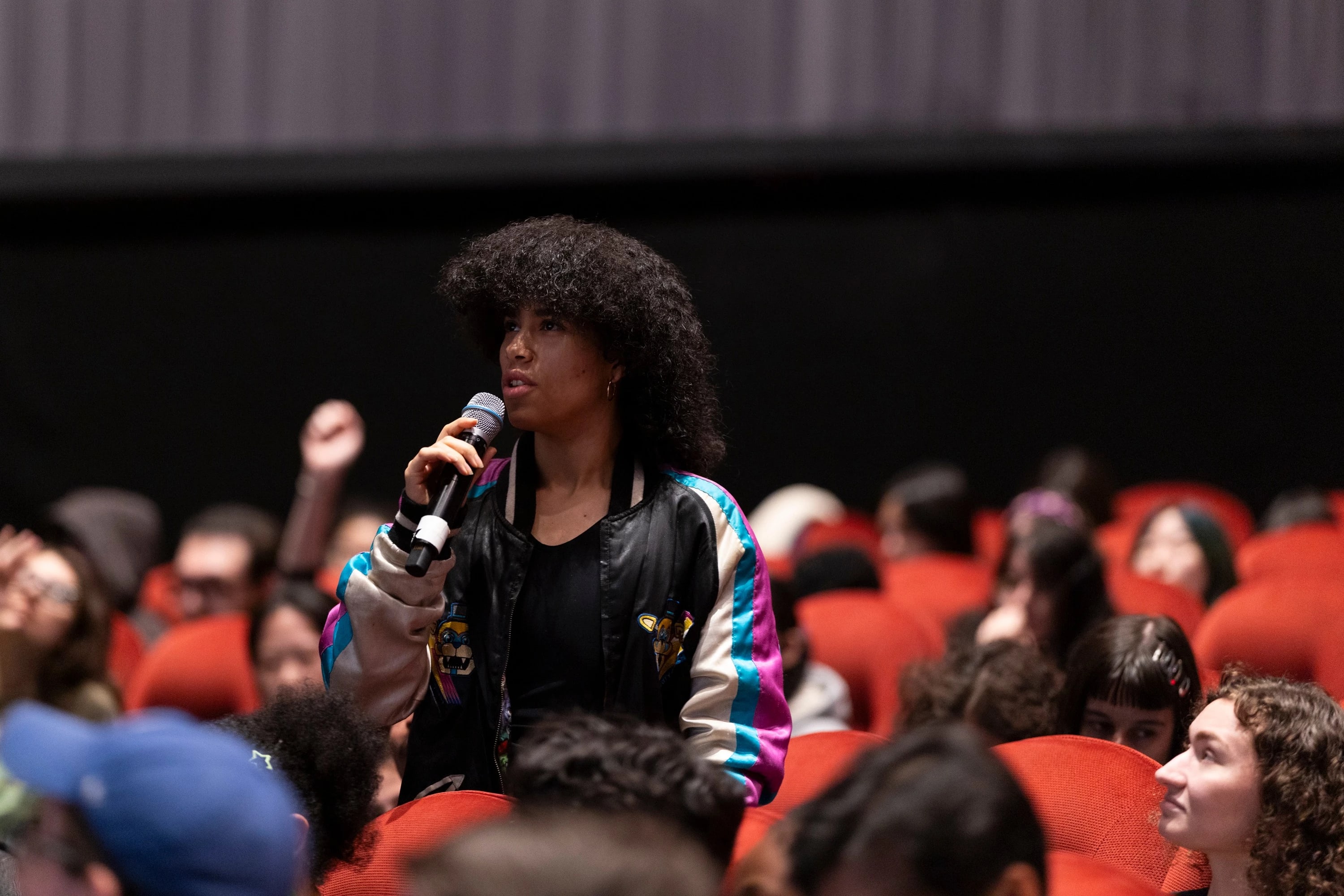 A person stands up among a seated audience, holding a microphone.