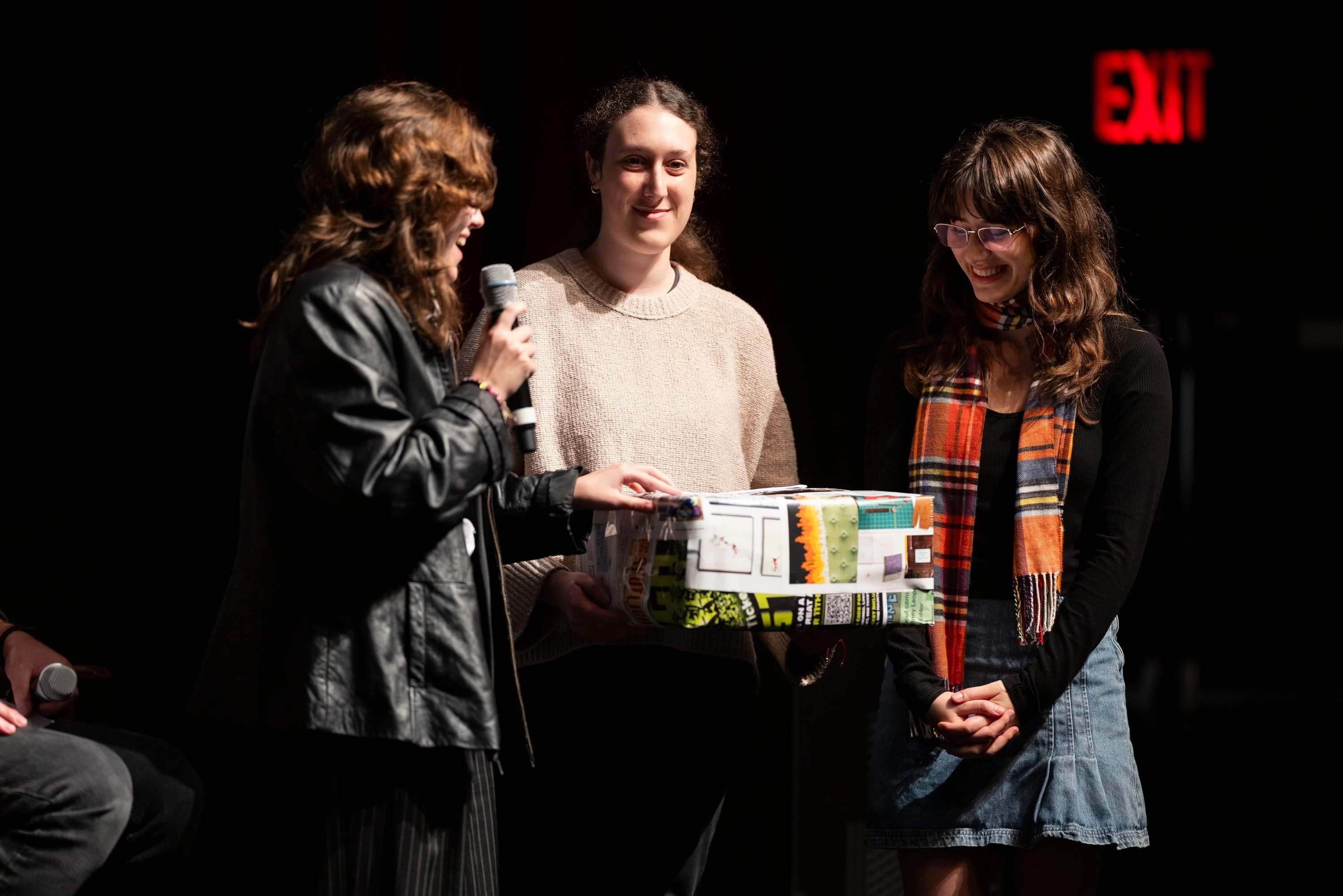 Three individuals stand on a stage holding a colorful box together.