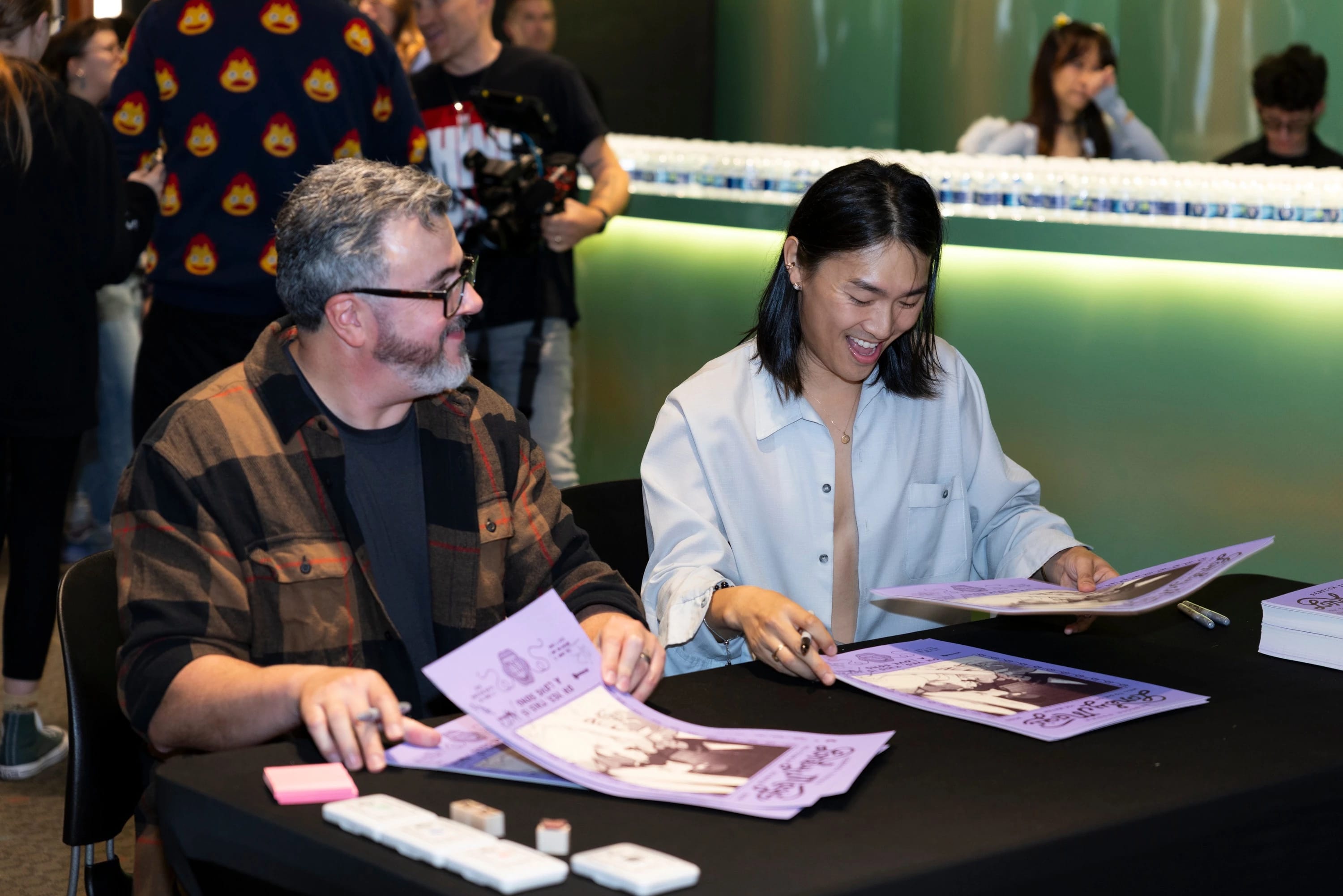 Two individuals smiling while sitting at a table, looking at posters and signing them.