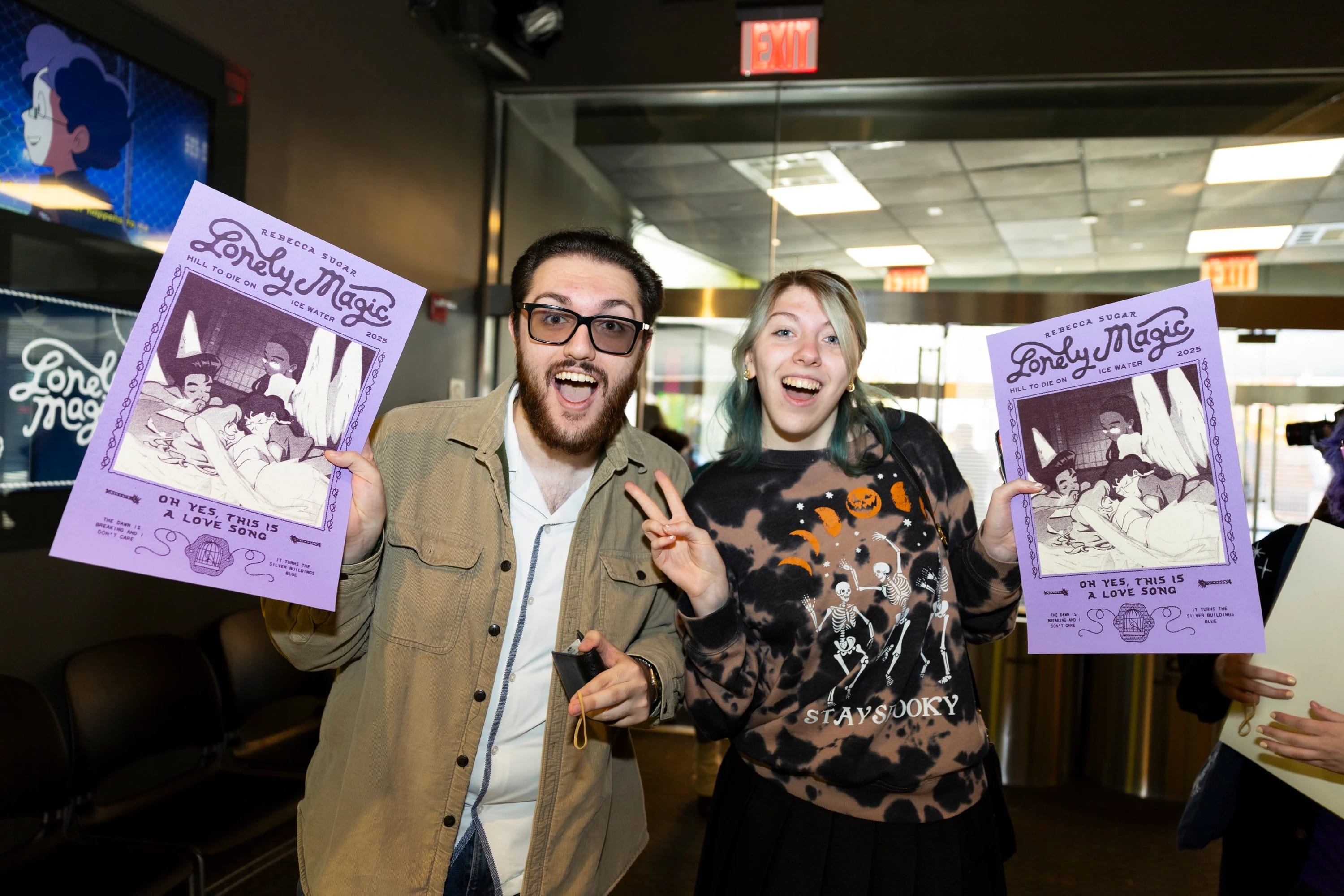 Two people holding purple posters, looking excited; one of them is raising a hand in a peace sign.