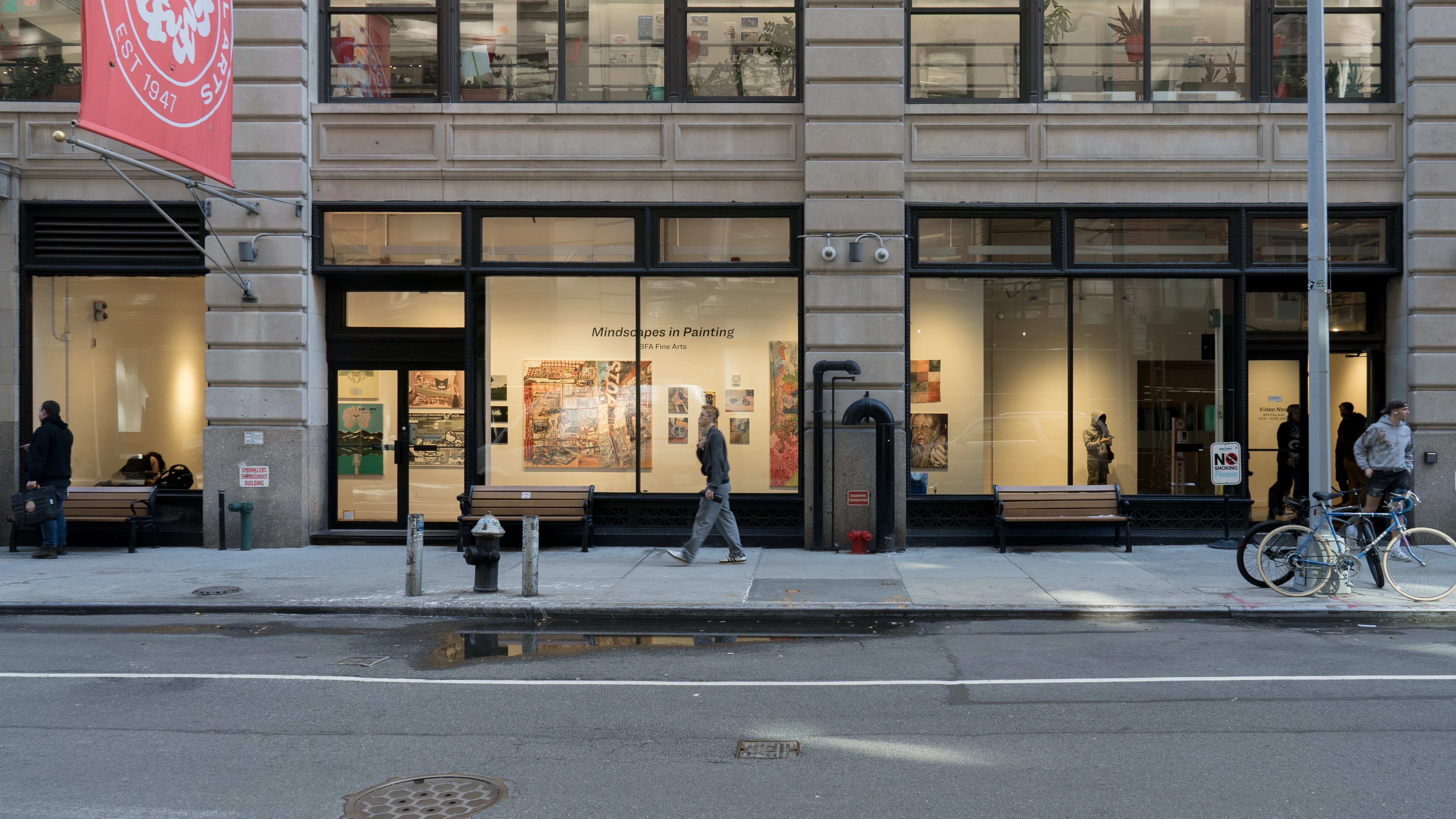 External view of an art gallery inside large picture windows, photographed from across a street, as pedestrians walk down the sidewalk running parallel. 