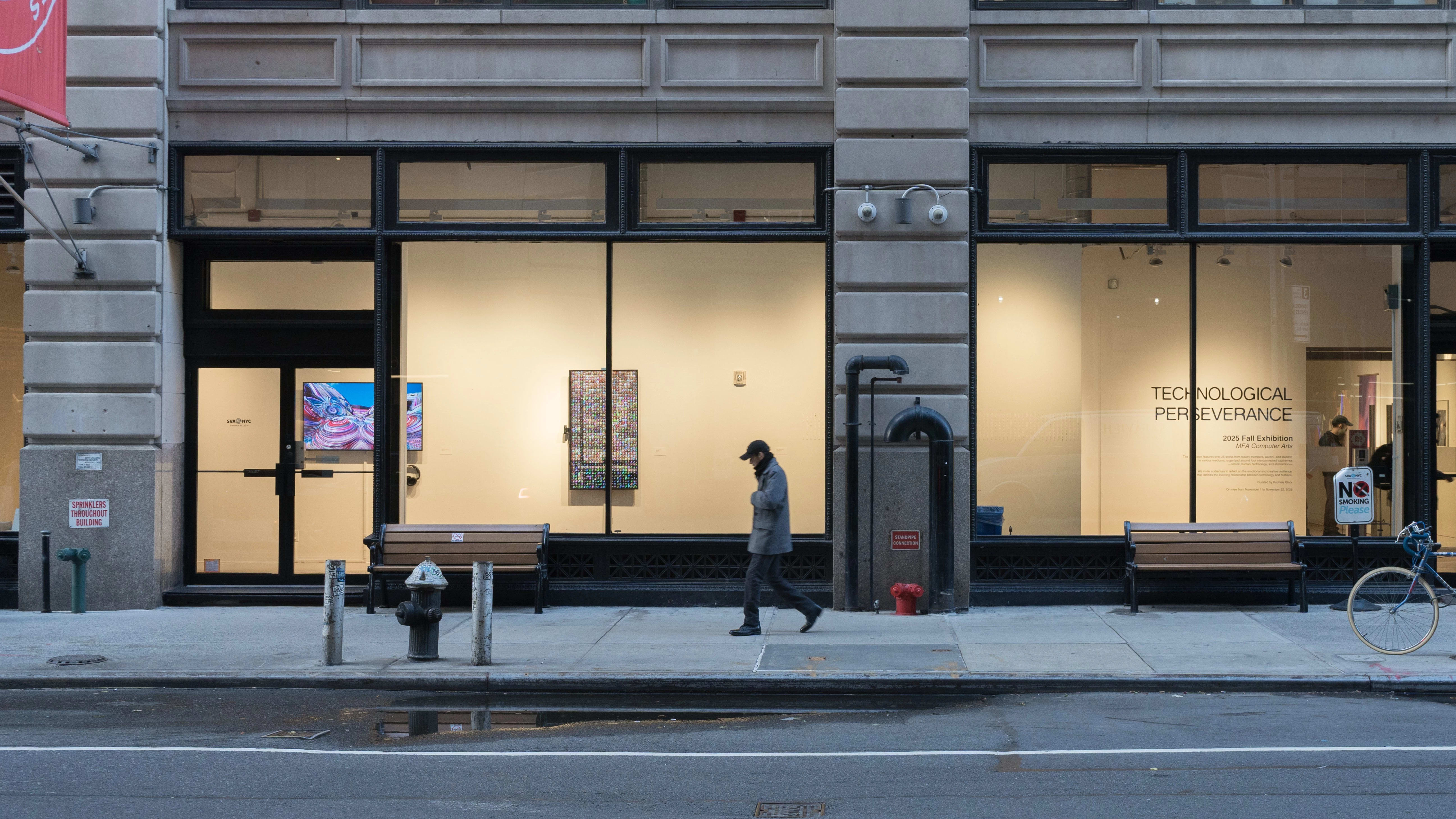From the outside, the SVA Flatiron Gallery is visible through the building's windows. Two monitors, one horizontal and one vertical, are on display in the hallway behind the windows. On the right side of the wall, vinyl text reads “Technological Perseverance.”