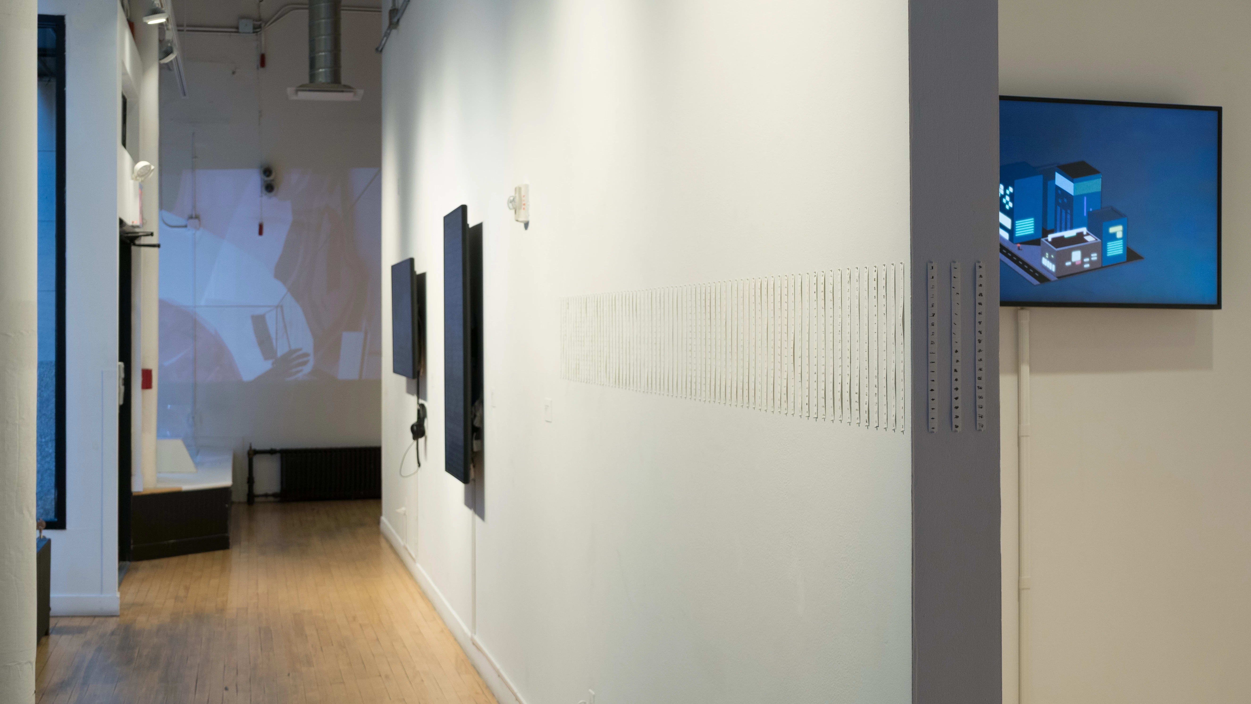 Looking down the hallway in front of the building windows, thin strips of paper are displayed in a sequence. Beyond them, there are two monitors on the wall and a projection at the end of the hall.