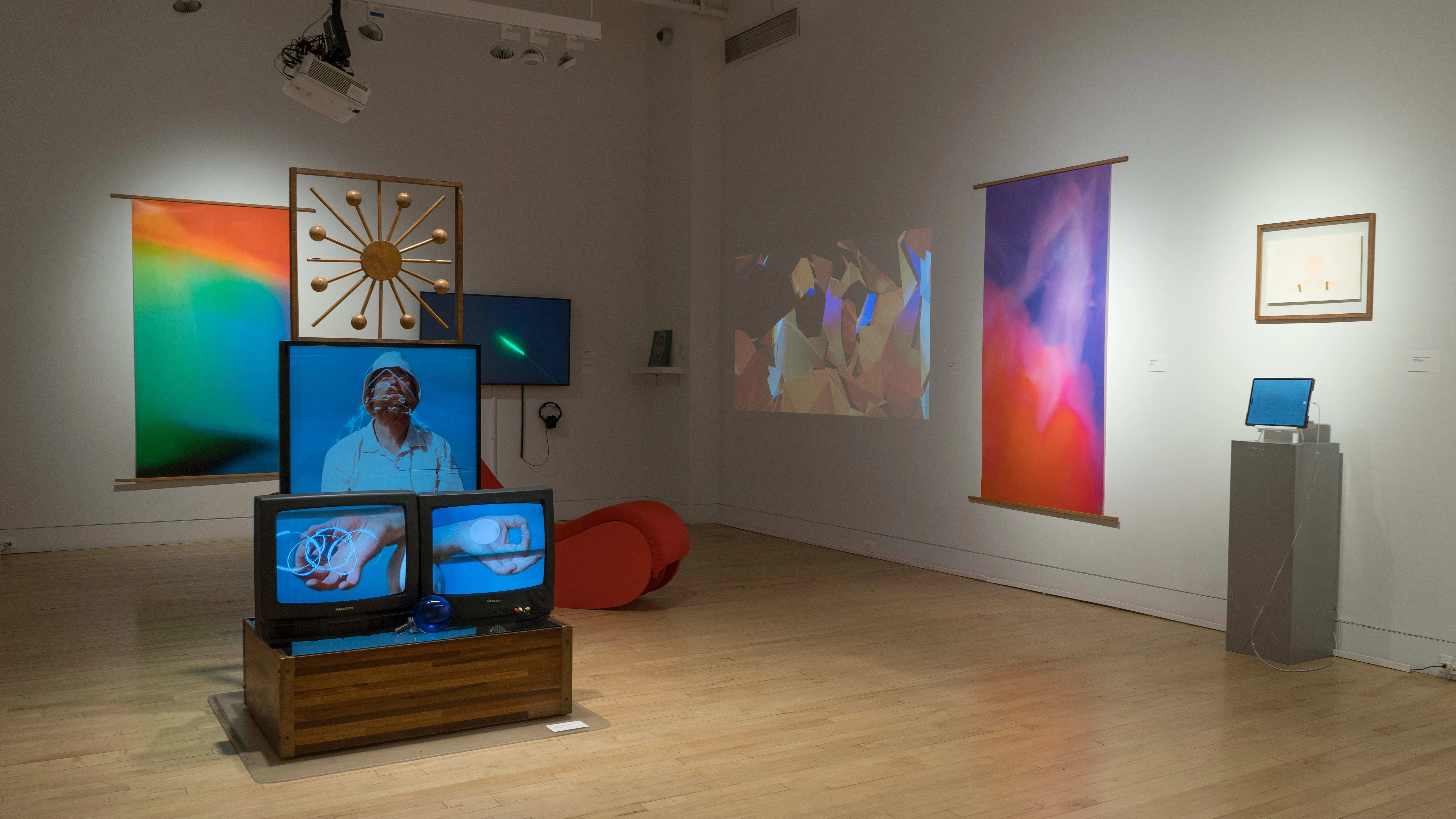 Inside the SVA Flatiron Gallery, colorful banners, monitors, and frames are hung on the walls. A projection is displayed on the right wall, while an installation featuring three televisions and a large, star-like wooden object occupies the center of the room.