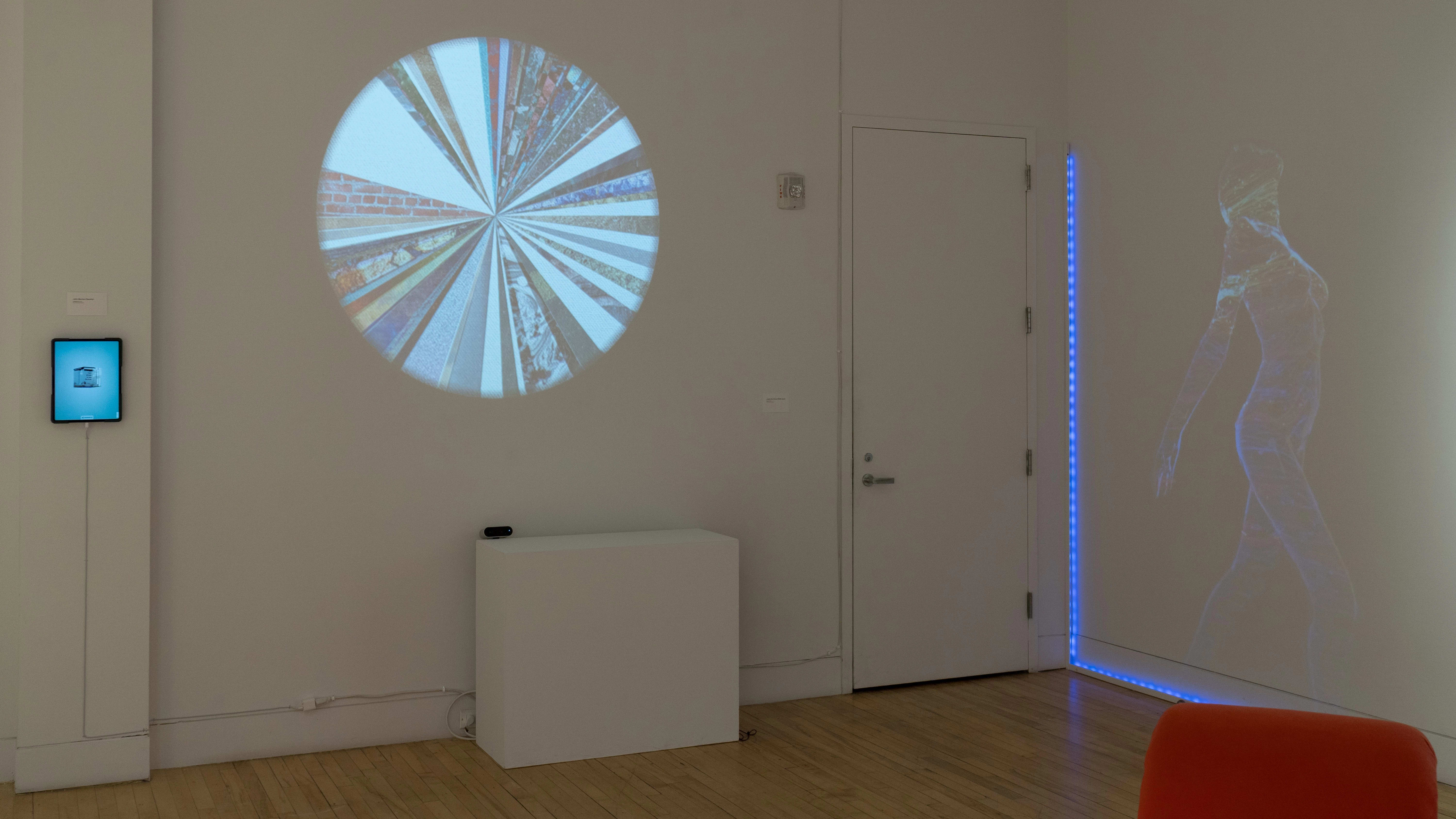In a corner of the gallery, a circle divided into triangular sections is projected on one wall. A woman’s body is faintly projected catty-corner to it. To the left, an iPad is mounted on the wall.