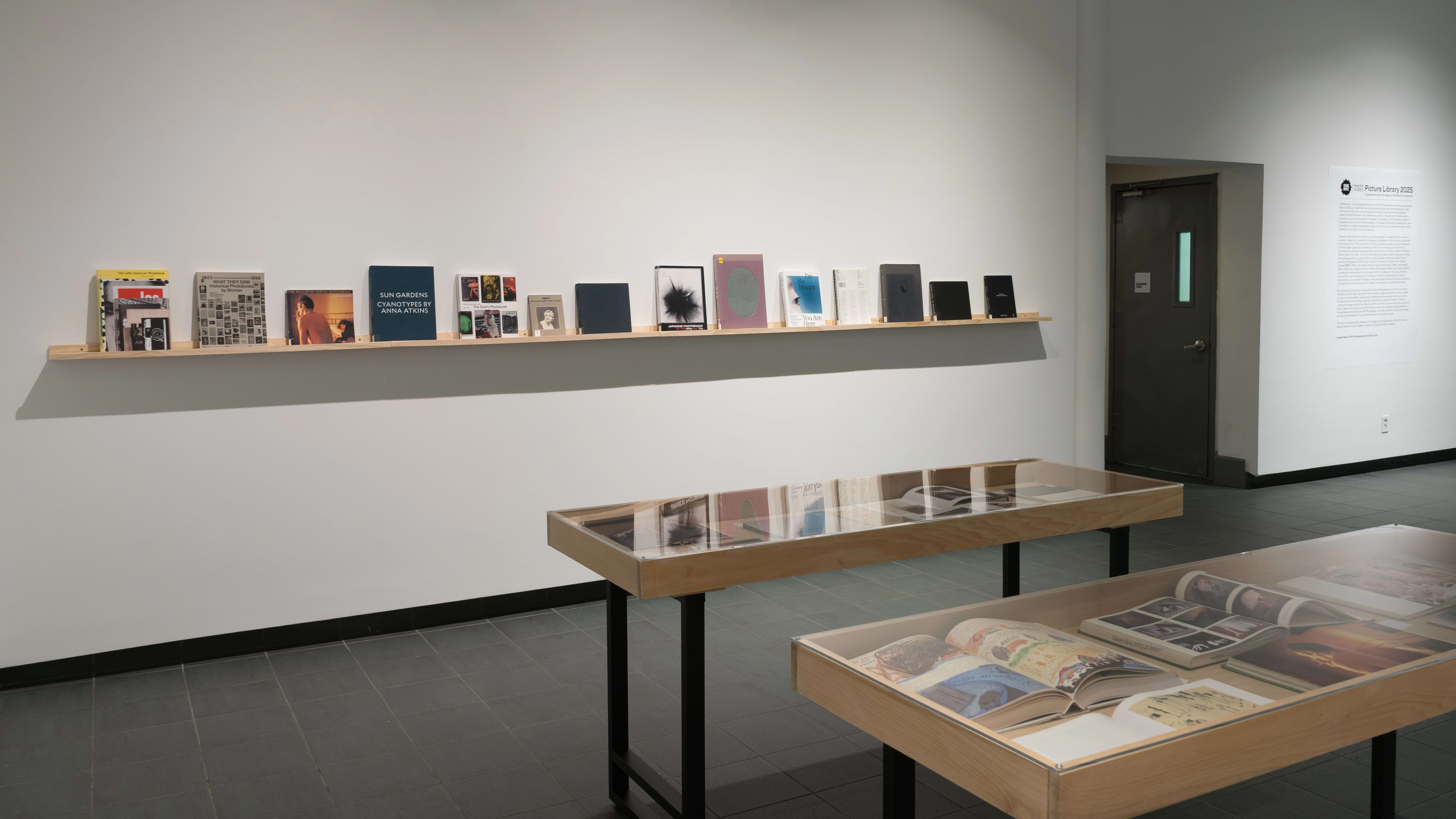 Two small vitrines stand in the center of the SVA Gramercy Gallery lobby. Behind them, one long wooden shelf runs down a wall. Many front-facing books of various sizes sit on the shelf.
