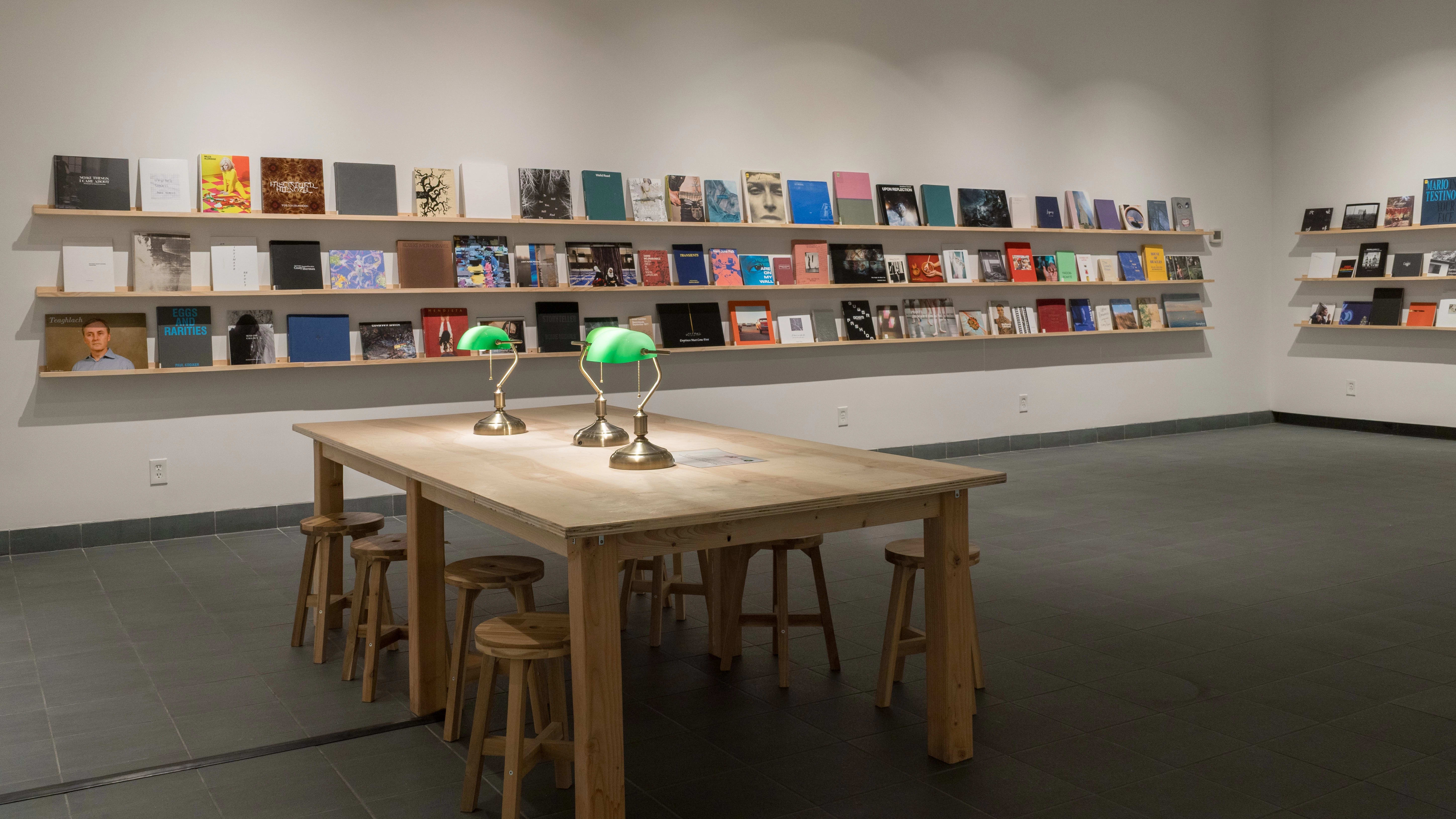 Each of the two visible walls is lined with three long, wooden shelves. Front-facing books fill all of the shelves. On the floor in front of them sit a large wooden table, a set of wooden stools, and three green table lamps.