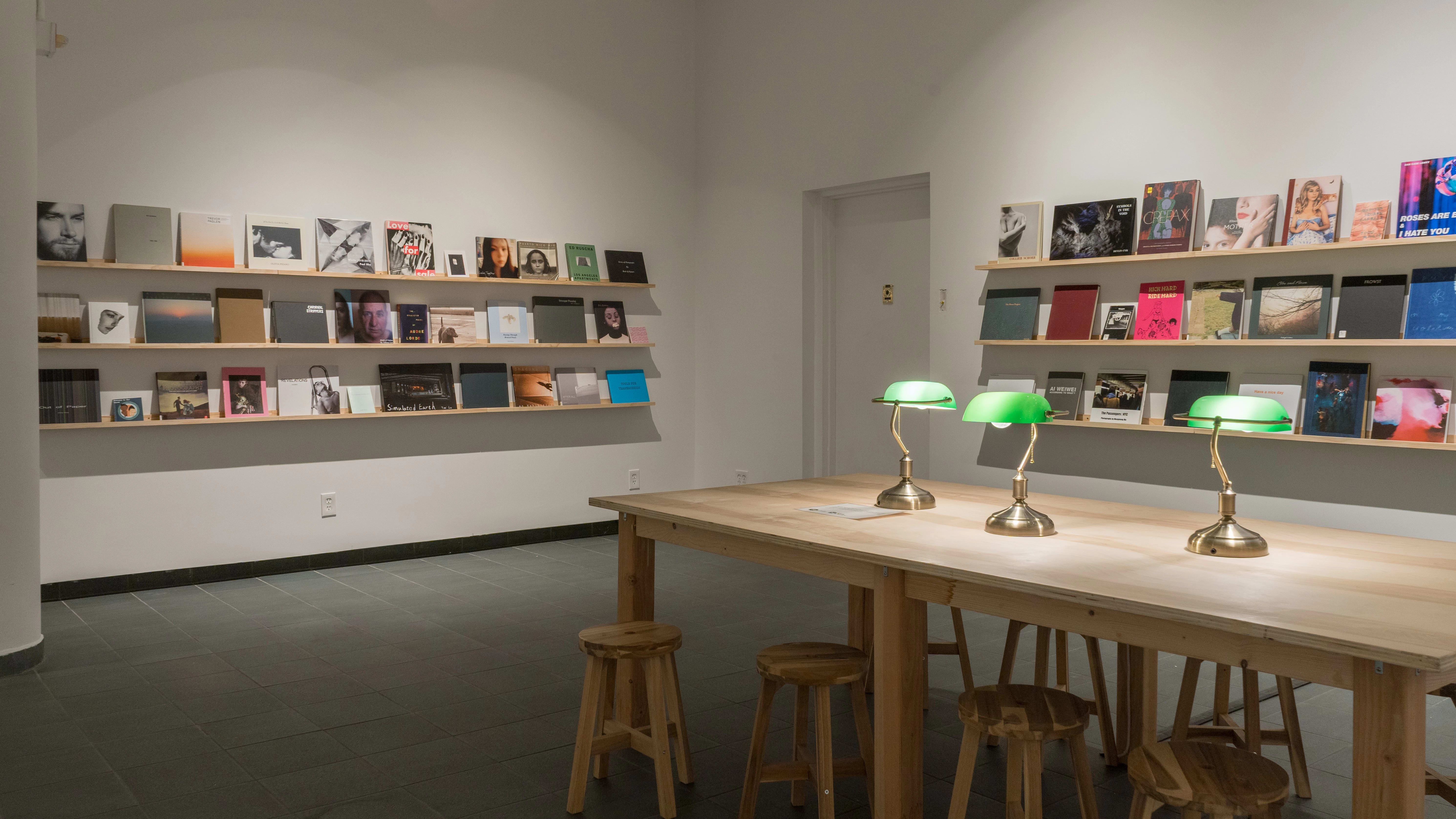 Each of the two visible walls is lined with three long, wooden shelves. Front-facing books of different sizes, colors, and designs fill all of the shelves. A large wooden table, accompanied by wooden stools and green table lamps, is set up in the center of the room.