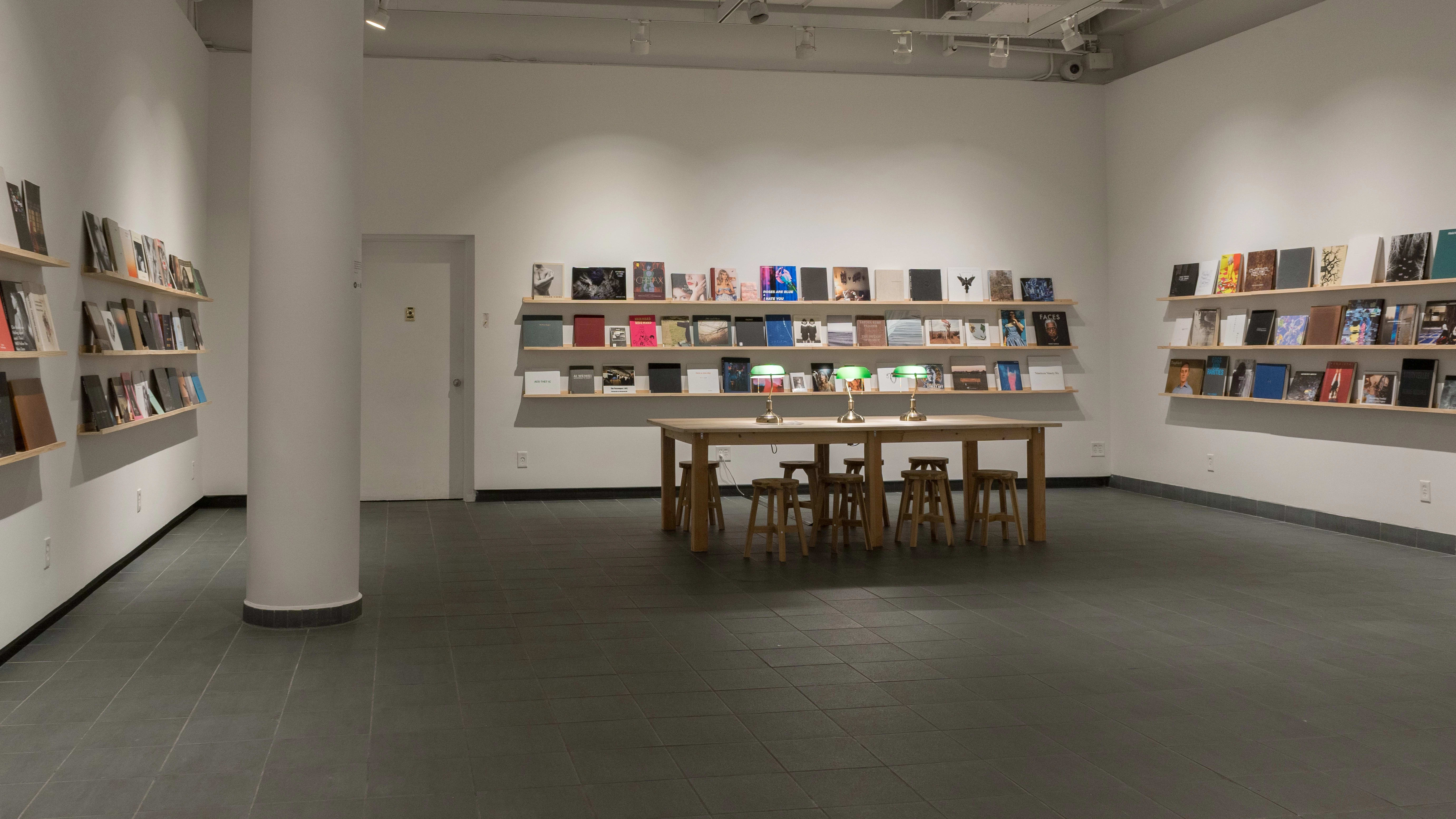 All of the walls in the gallery are lined with long, wooden shelves. Front-facing books of different sizes, colors, and designs fill all of the shelves. A large wooden table, accompanied by wooden stools and green table lamps, is set up in the center of the room.