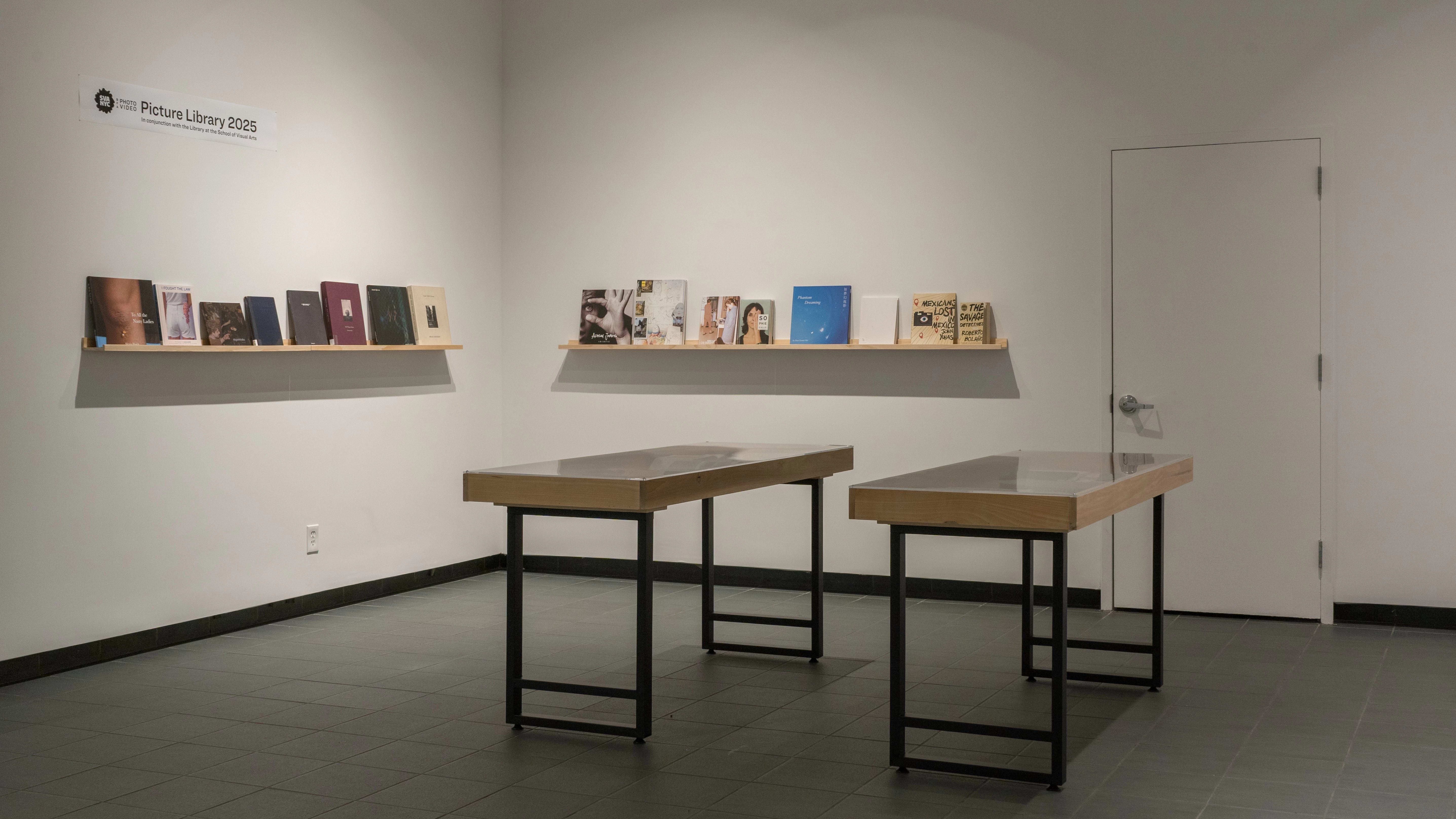 Two small vitrines stand in the center of the SVA Gramercy Gallery lobby. Behind them, two walls each have a single wooden shelf holding front-facing books of various sizes. A small sign above the shelves reads “Picture Library 2025.”