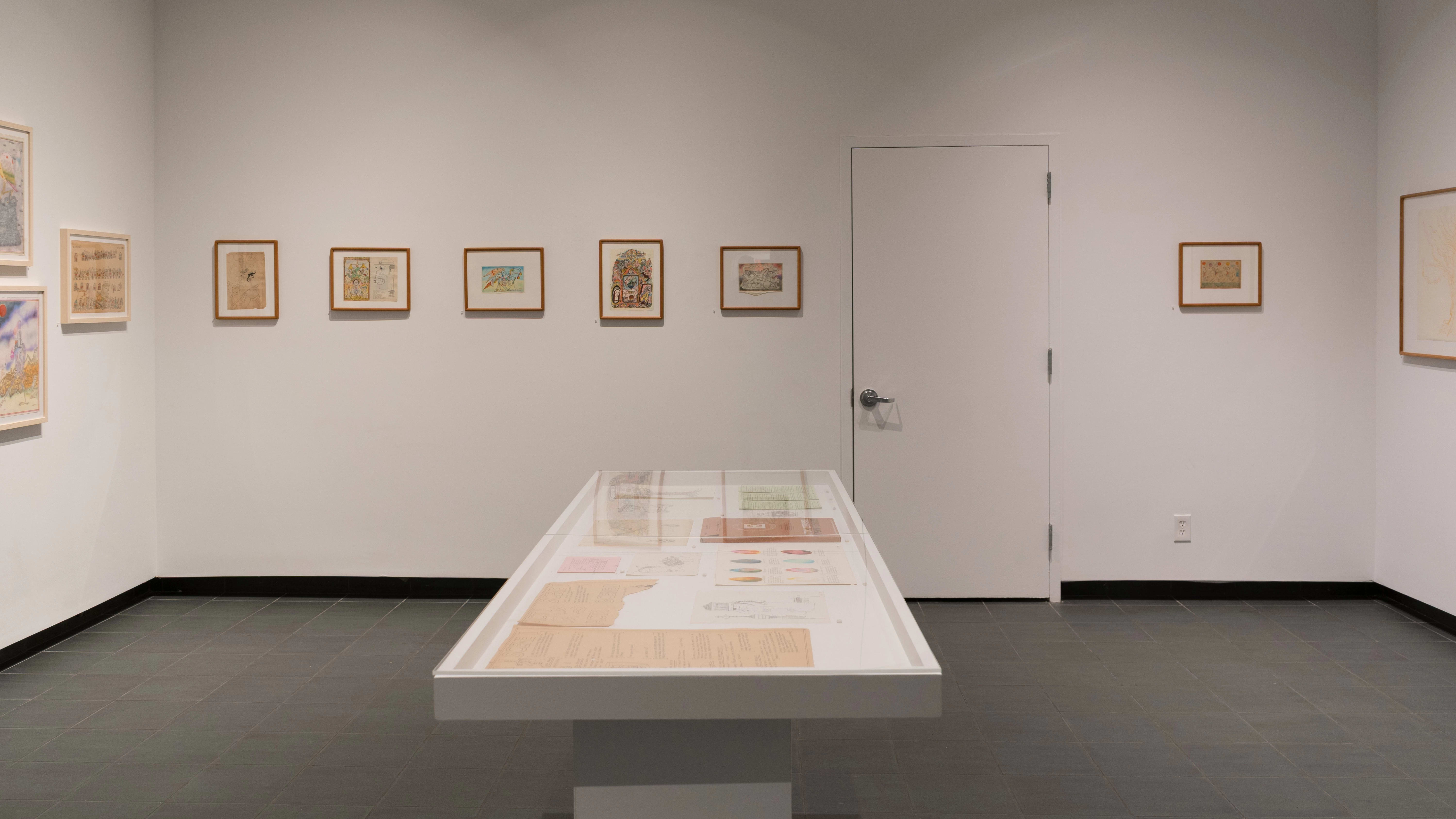 On one wall, small illustrations are framed in wooden frames with white mats. Some are oriented vertically, while others are oriented horizontally. A vitrine, in front of the walls, holds loose papers.