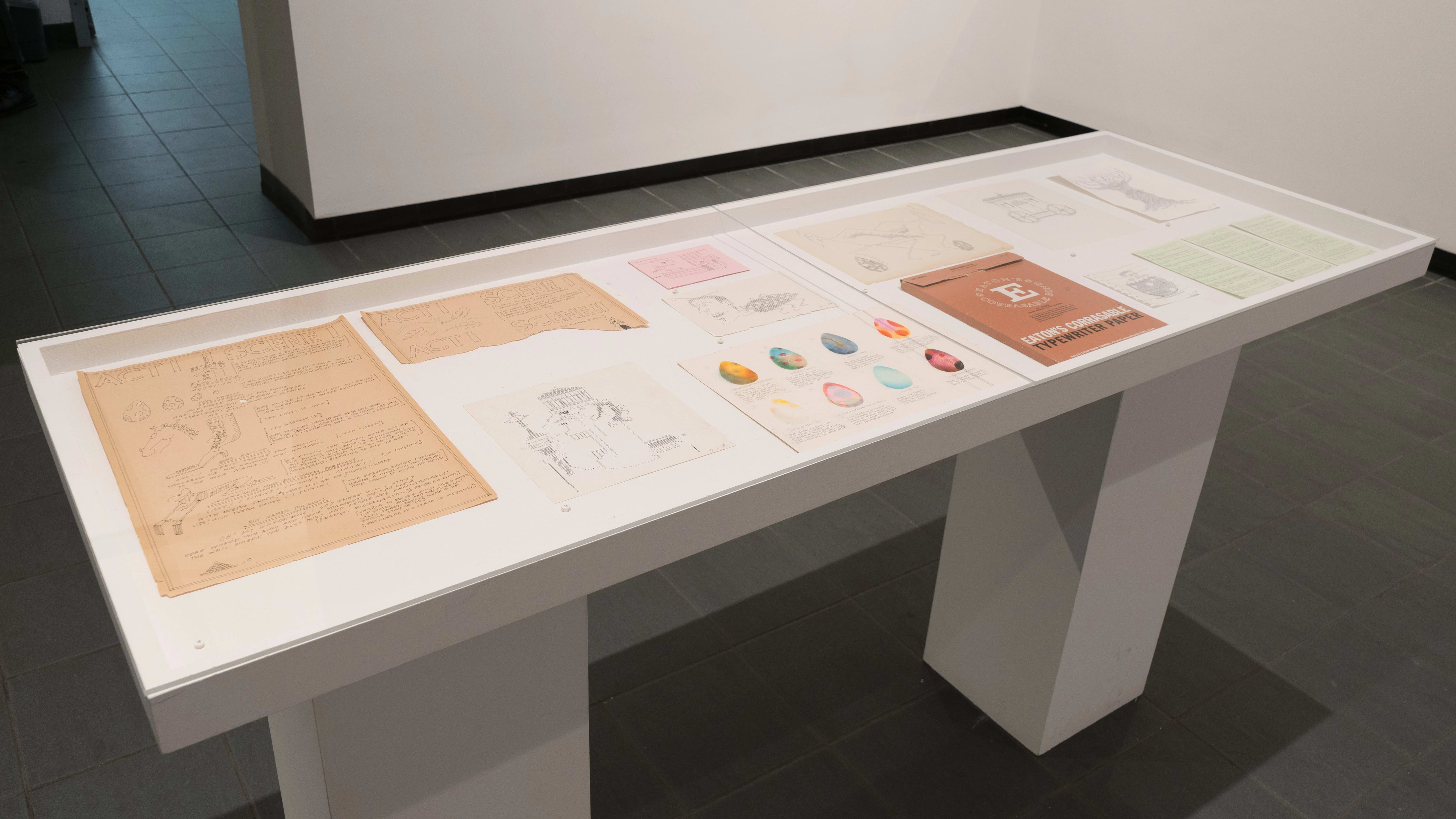 Looking down into a vitrine, papers of various sizes and colors are laid out. On some papers, there are sketches of fantastical characters or objects.