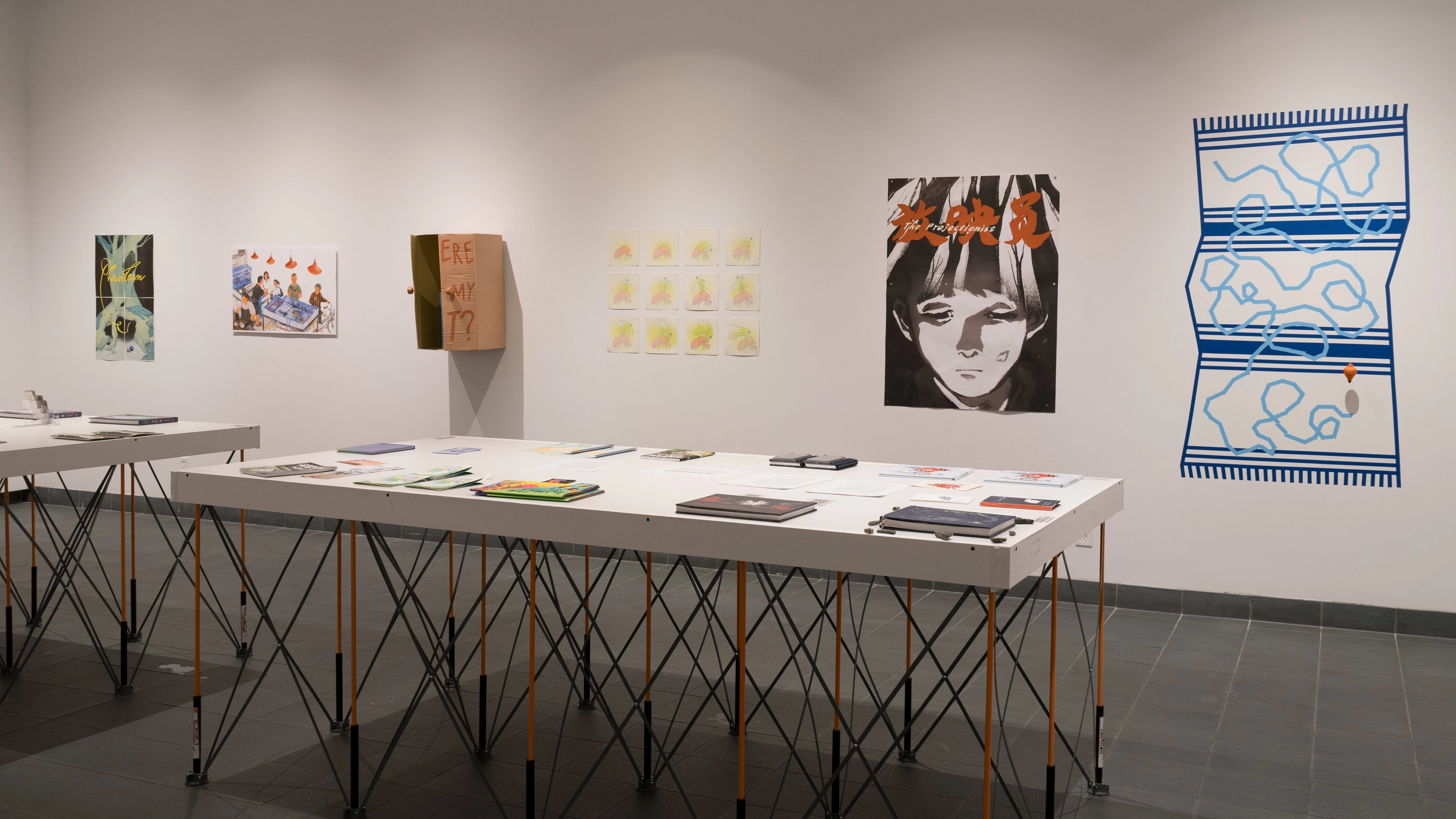 In the main room of the SVA Gramercy Gallery, two white tables displaying several books are set up near the center of the floor. Behind them, prints, vinyl, and sculptures are displayed on the wall.