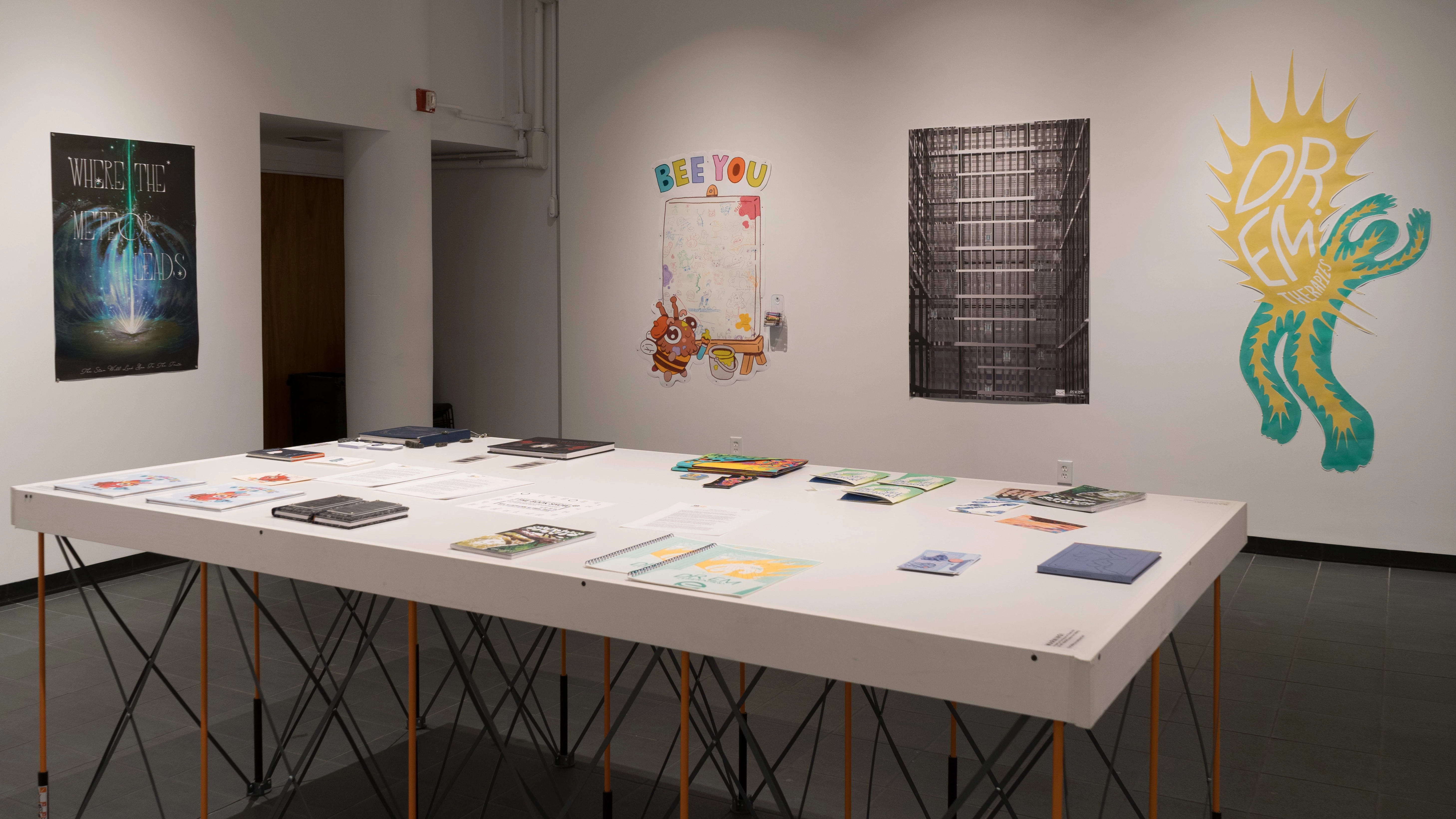 Many books lay flat on a white table near the entrance to the room. Beyond the table, prints and cut-paper artworks are displayed on the wall.