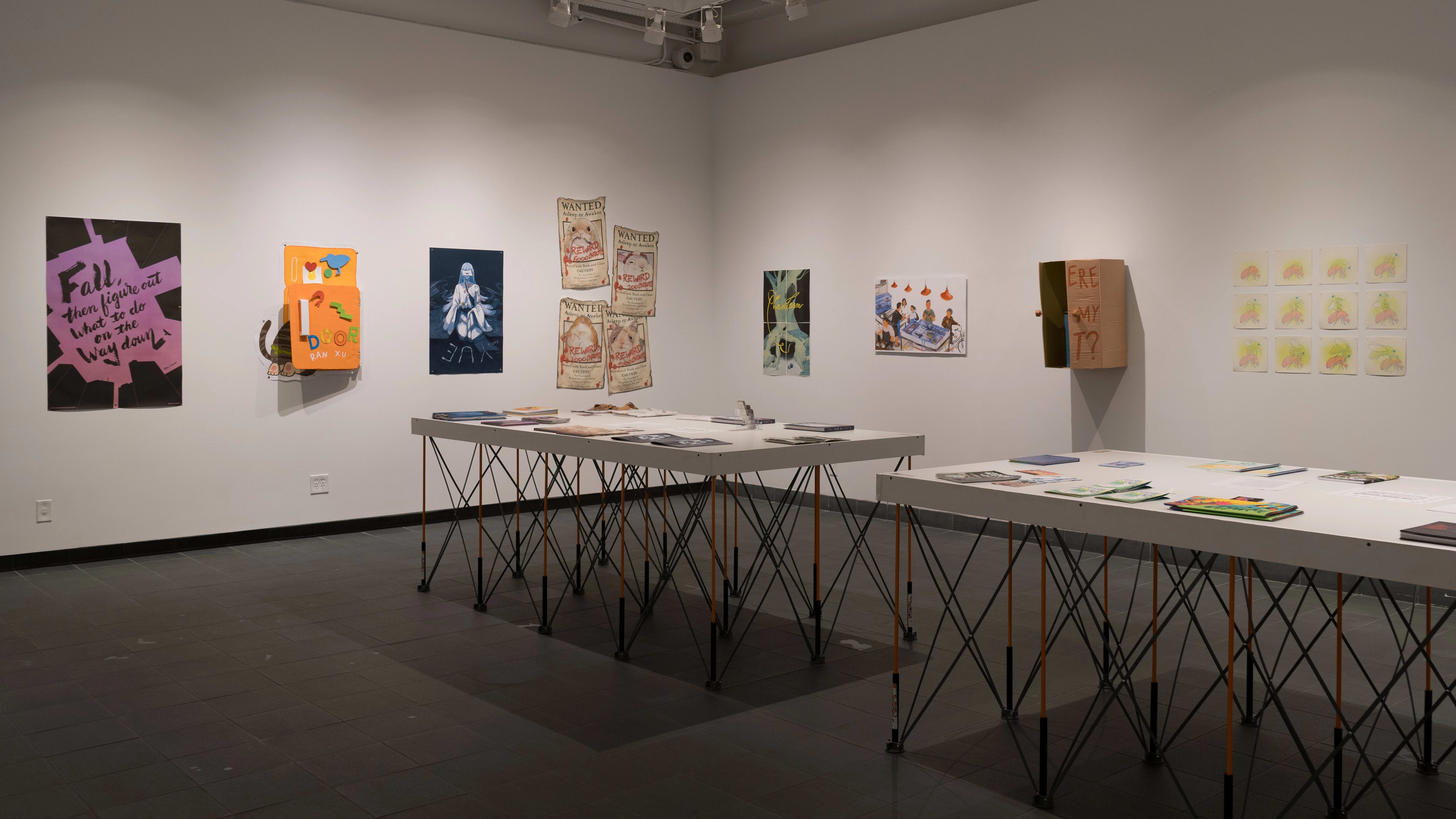 Two tables in the gallery display many books, laid flat. Prints, posters, layered papers, and cardboard sculptures are hung on the surrounding walls.