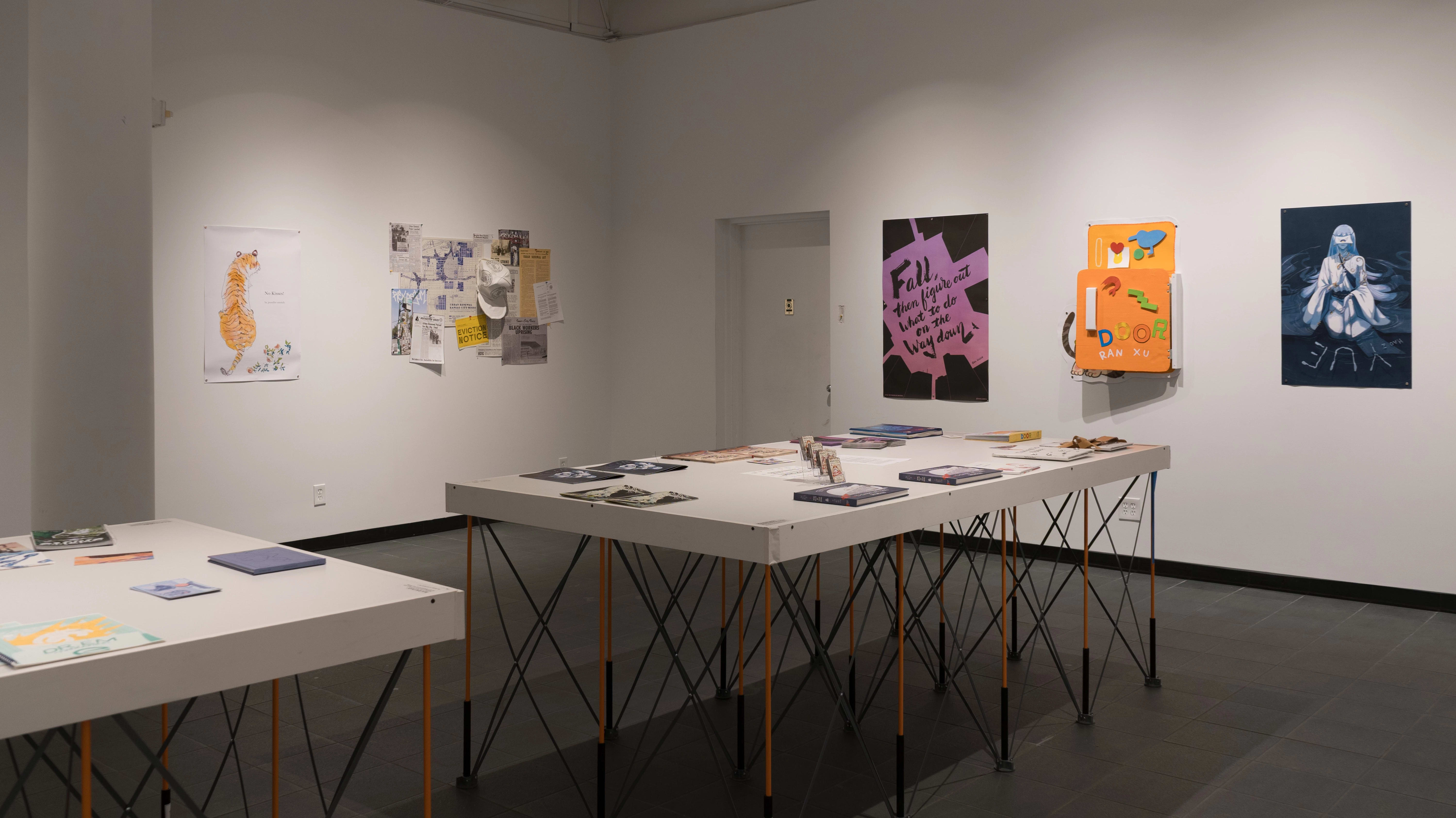 Two tables in the gallery display many books, laid flat. Prints, posters, layered papers, and cardboard sculptures are hung on the surrounding walls. The works vary in color, size, style, and content.