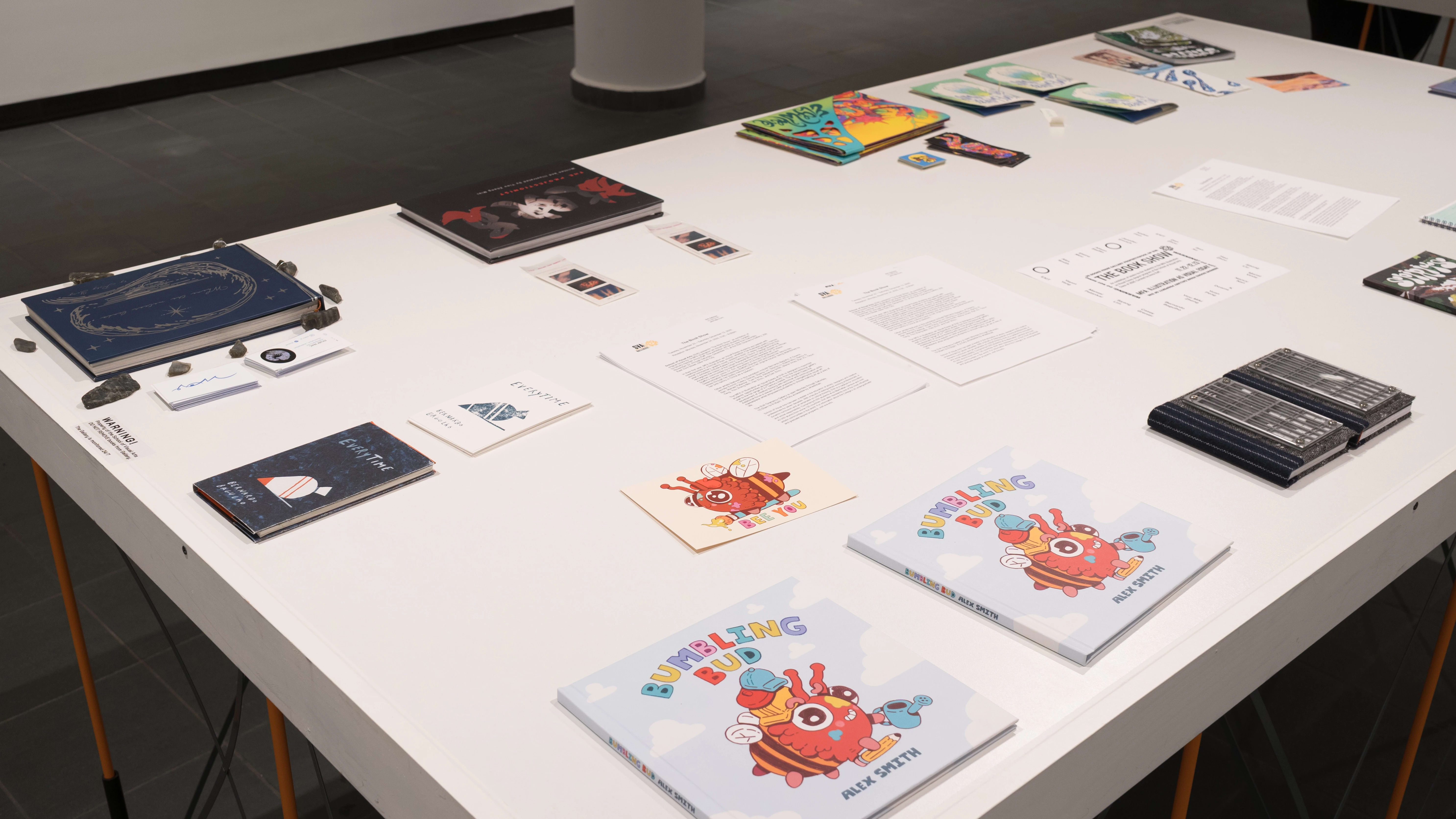 Press releases and a gallery map lay in the center of a table in the gallery. Many different books are arranged around the papers. The nearest book is titled “Bumbling Bud” and depicts a smiling red and orange bumblebee-like creature.