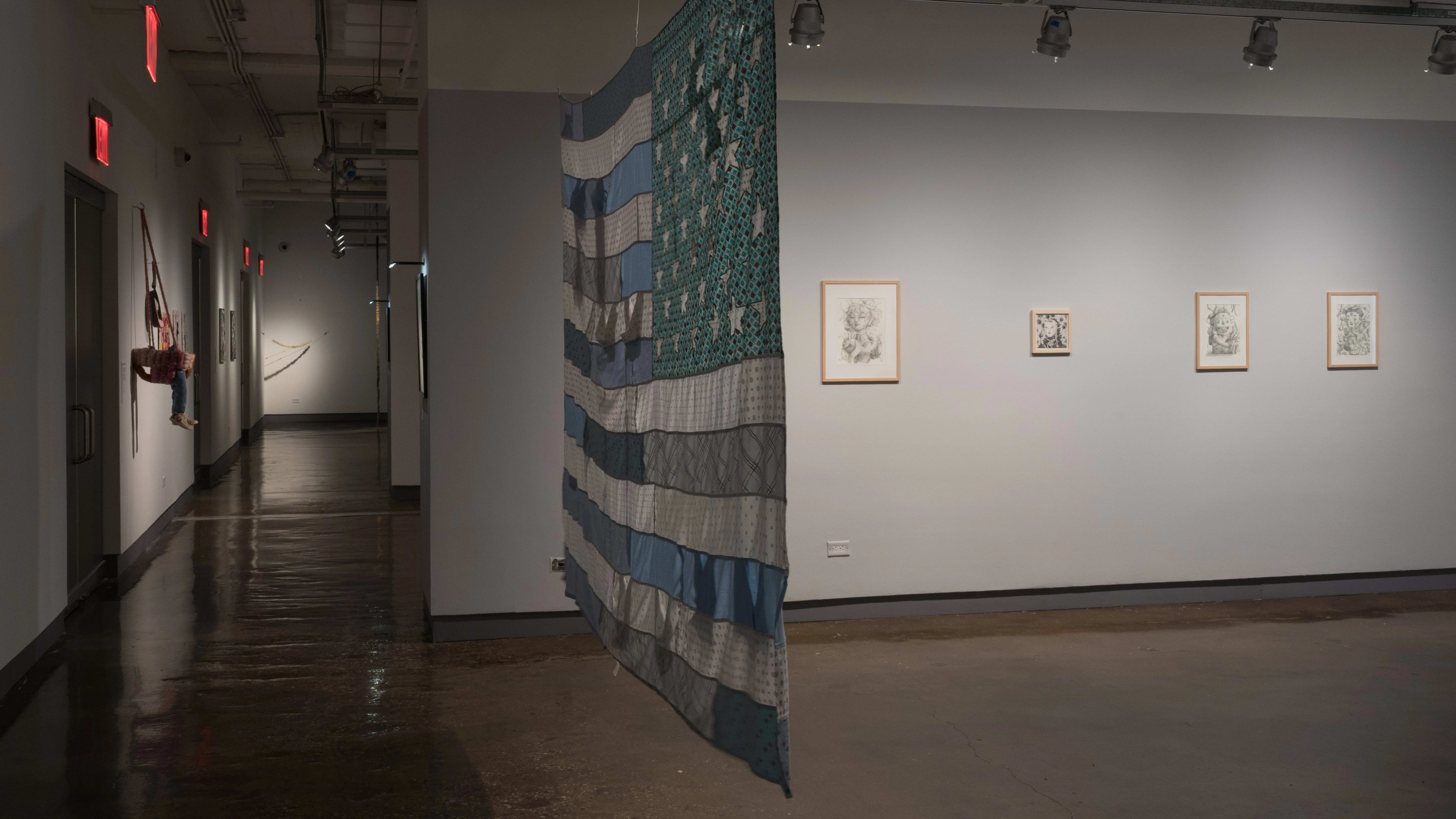 A hallway of the SVA Chelsea Gallery behind a flag artwork, with several other artworks on the walls to the left and right.