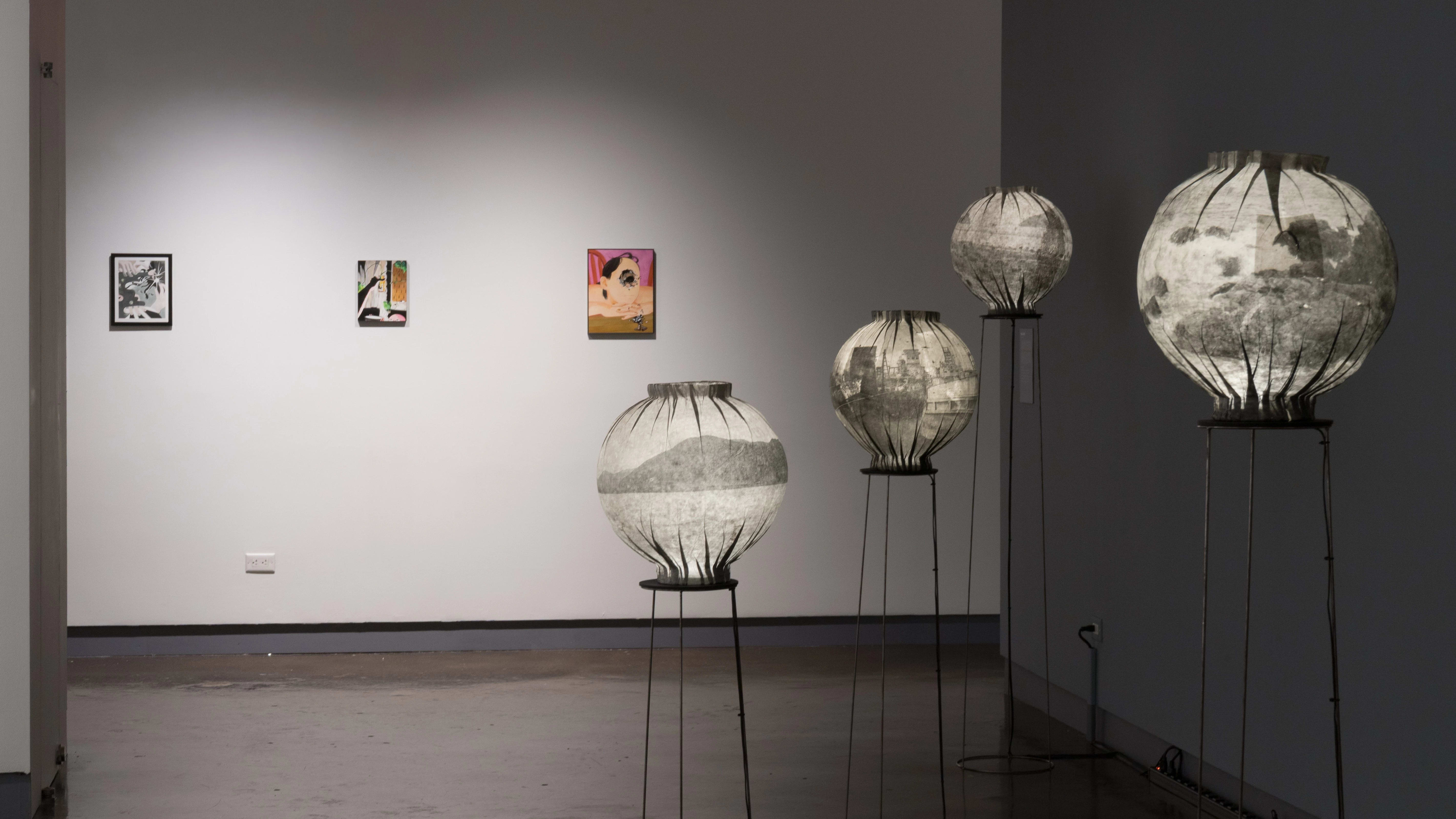 An image looking down the third gallery with paintings on the far wall, and paper lanterns on wire pedestals in front.