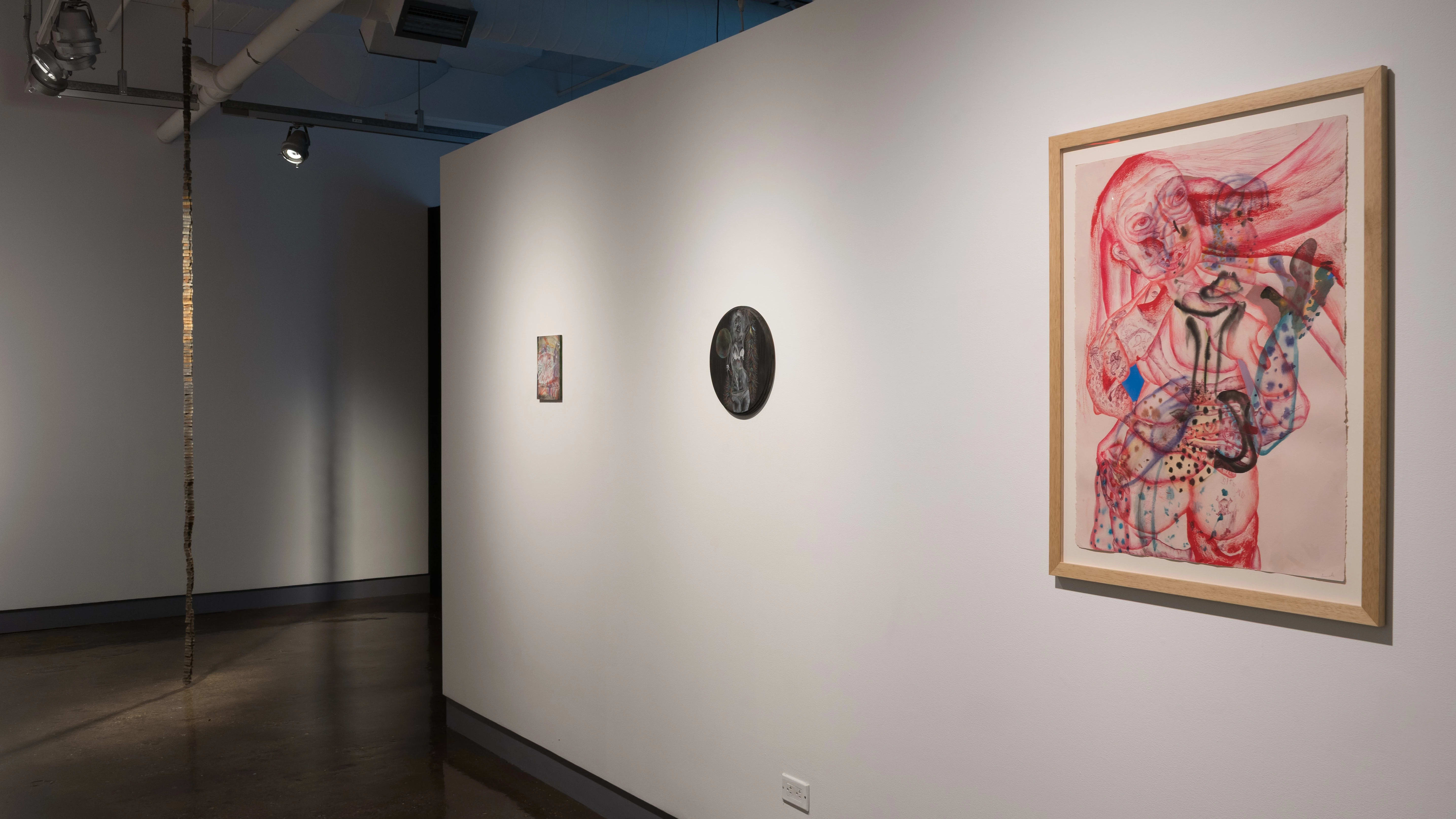 Three pastel drawings of various sizes and colors hang on a wall in the hallway of the gallery. A sculpture in a long strand hangs from the ceiling ahead.