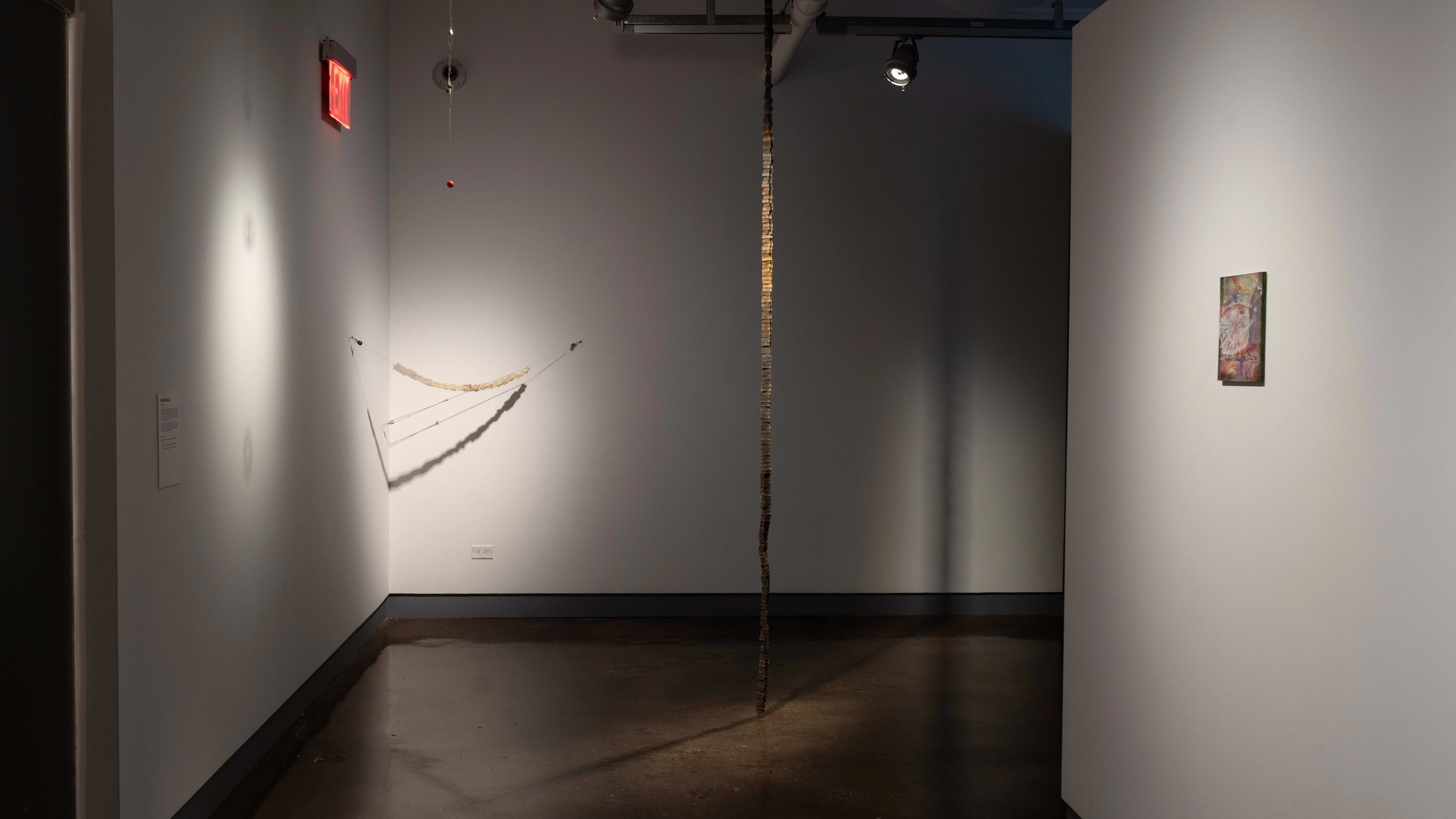 A corner of the gallery where a sculptural piece hangs from the ceiling to the floor, and another hangs diagonally across the wall.