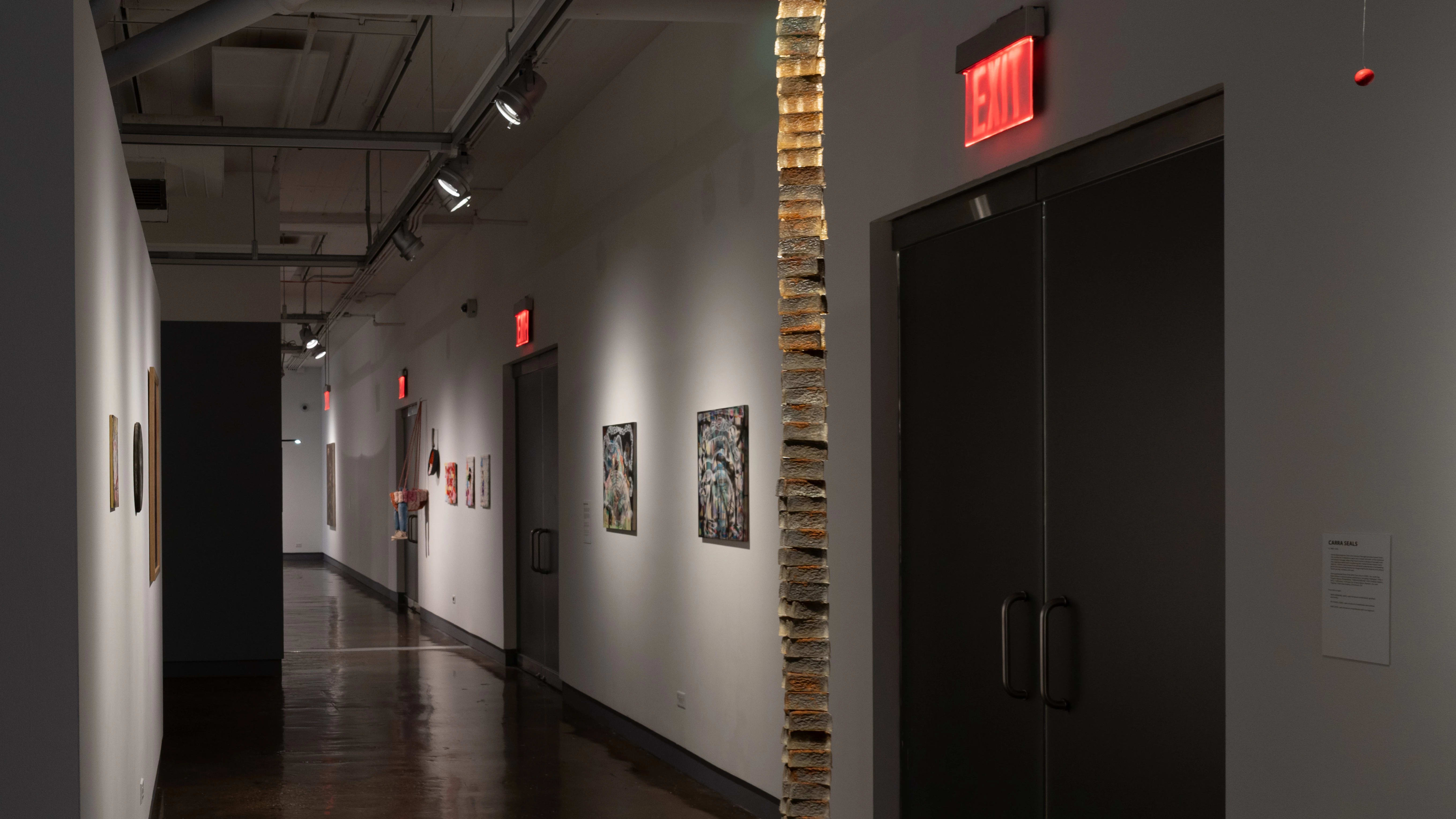 A hallway in SVA Chelsea Gallery, with artwork and sculptural pieces hanging on the left and right walls.