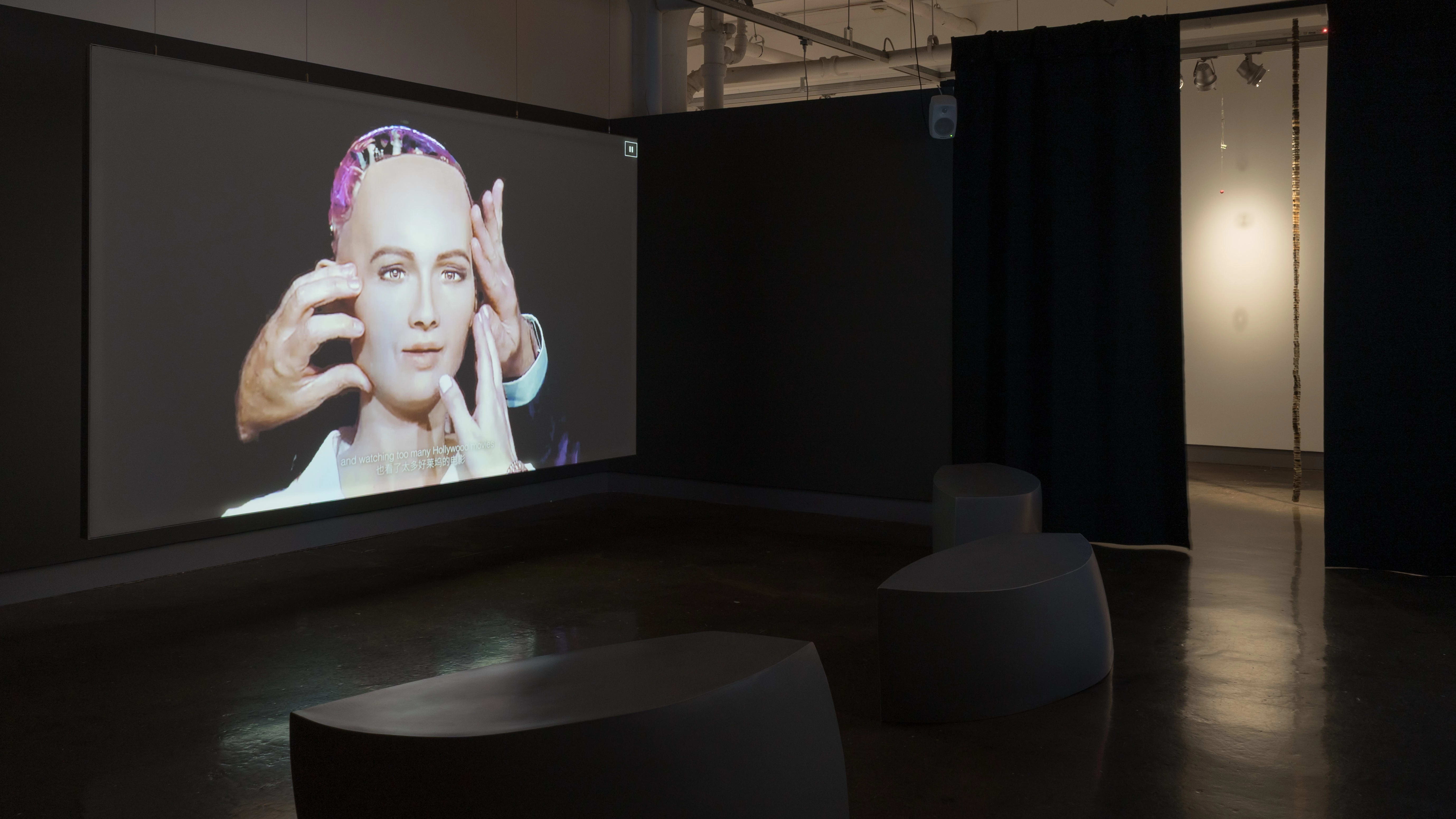 A large screen surrounded by seats, playing a video of a robot with a human-like face.