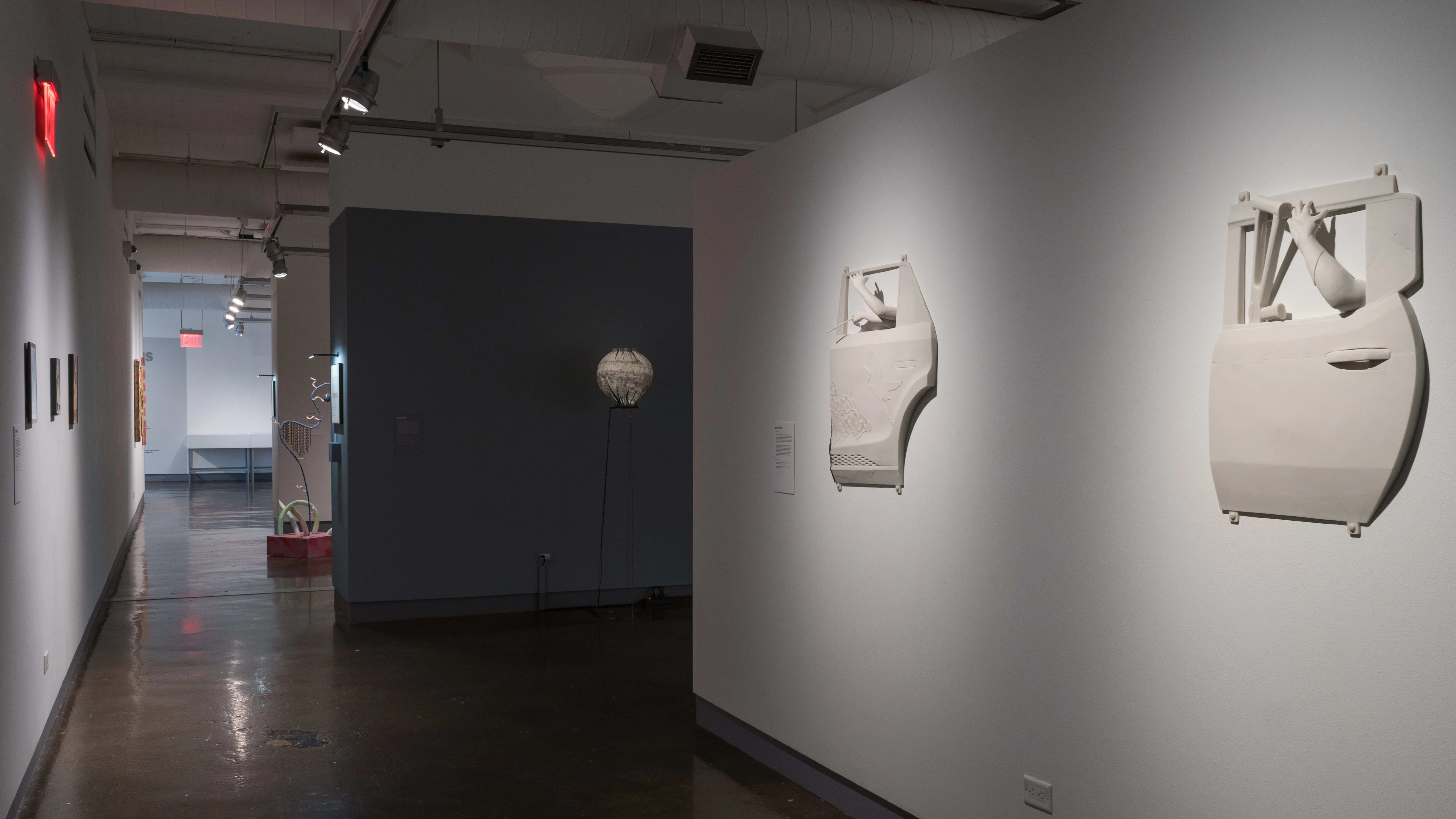 In a hallway of the SVA Chelsea Gallery, flat works hang from the wall on the left side, and sculptural works are seen in the third gallery.