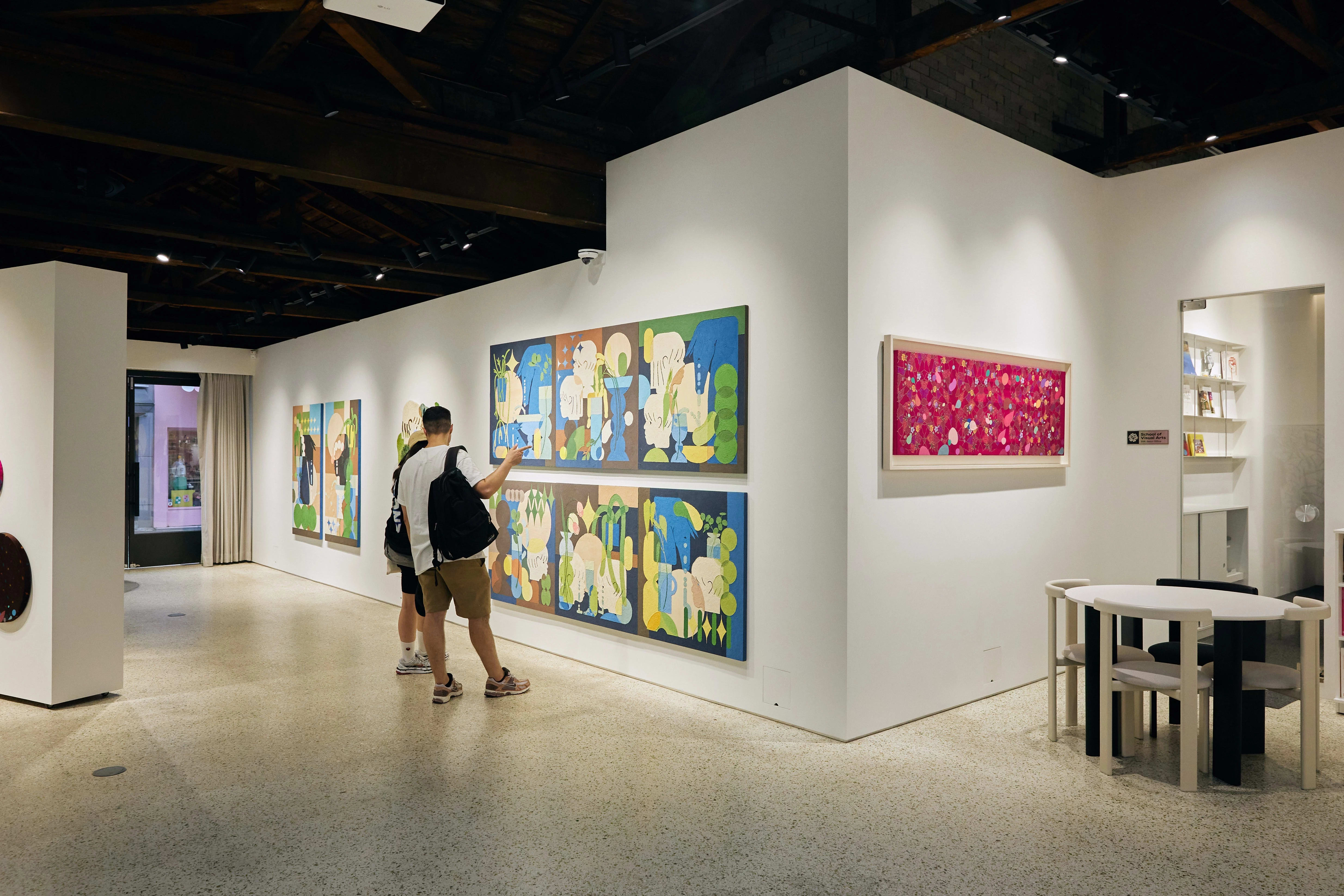 Gallery scene with two visitors viewing large, colorful paintings of abstract botanical forms and figures. A vivid pink artwork hangs nearby, and a reading area with modern white-and-black furniture is visible in the corner.
