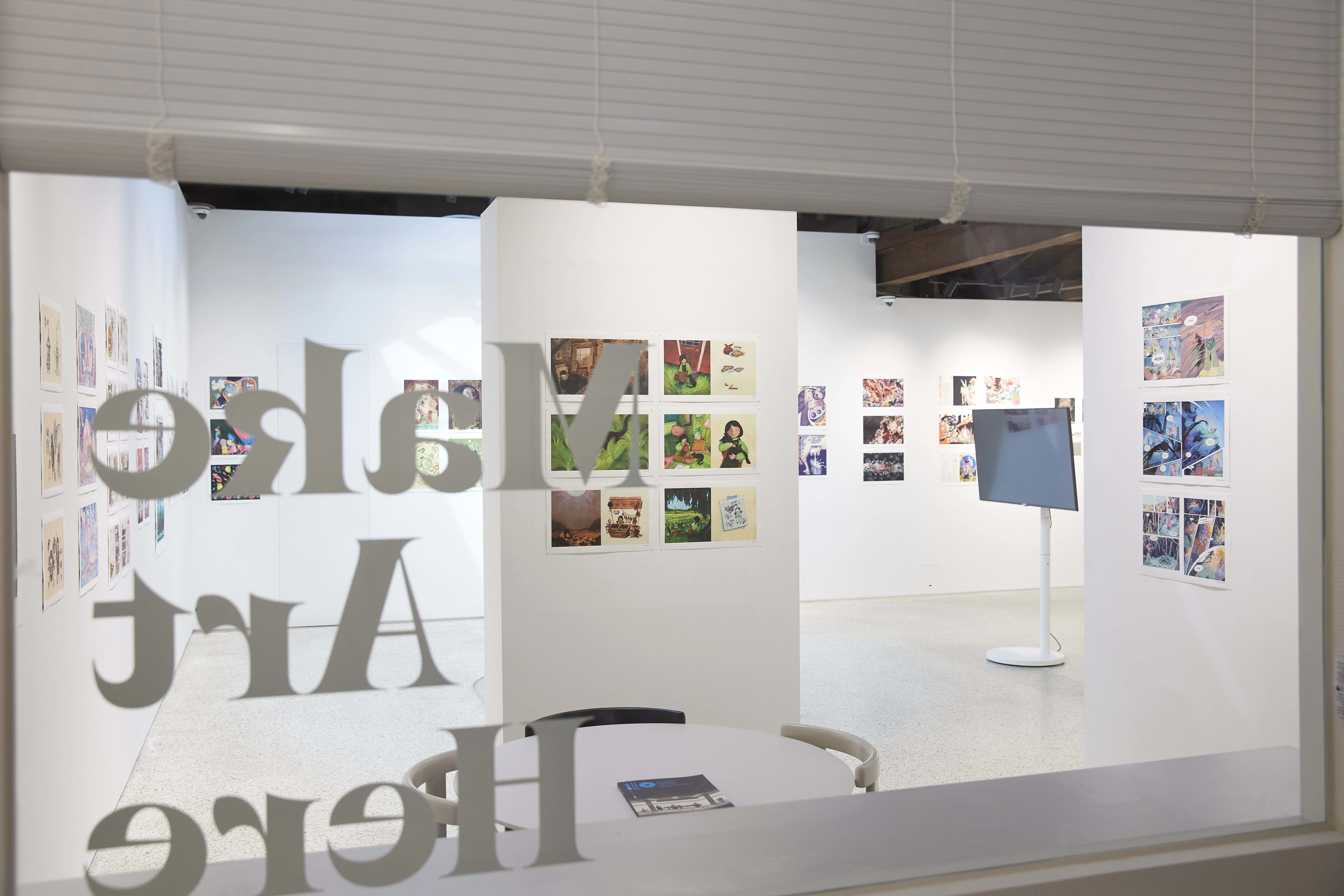 View of a bright gallery interior seen through a window with the words “Make Art Here” in reverse on the glass. The exhibition features colorful illustration and comic-style artworks arranged in neat grids on white walls. A digital display monitor stands on a white pedestal at the center, and a round table with a brochure is visible in the foreground.