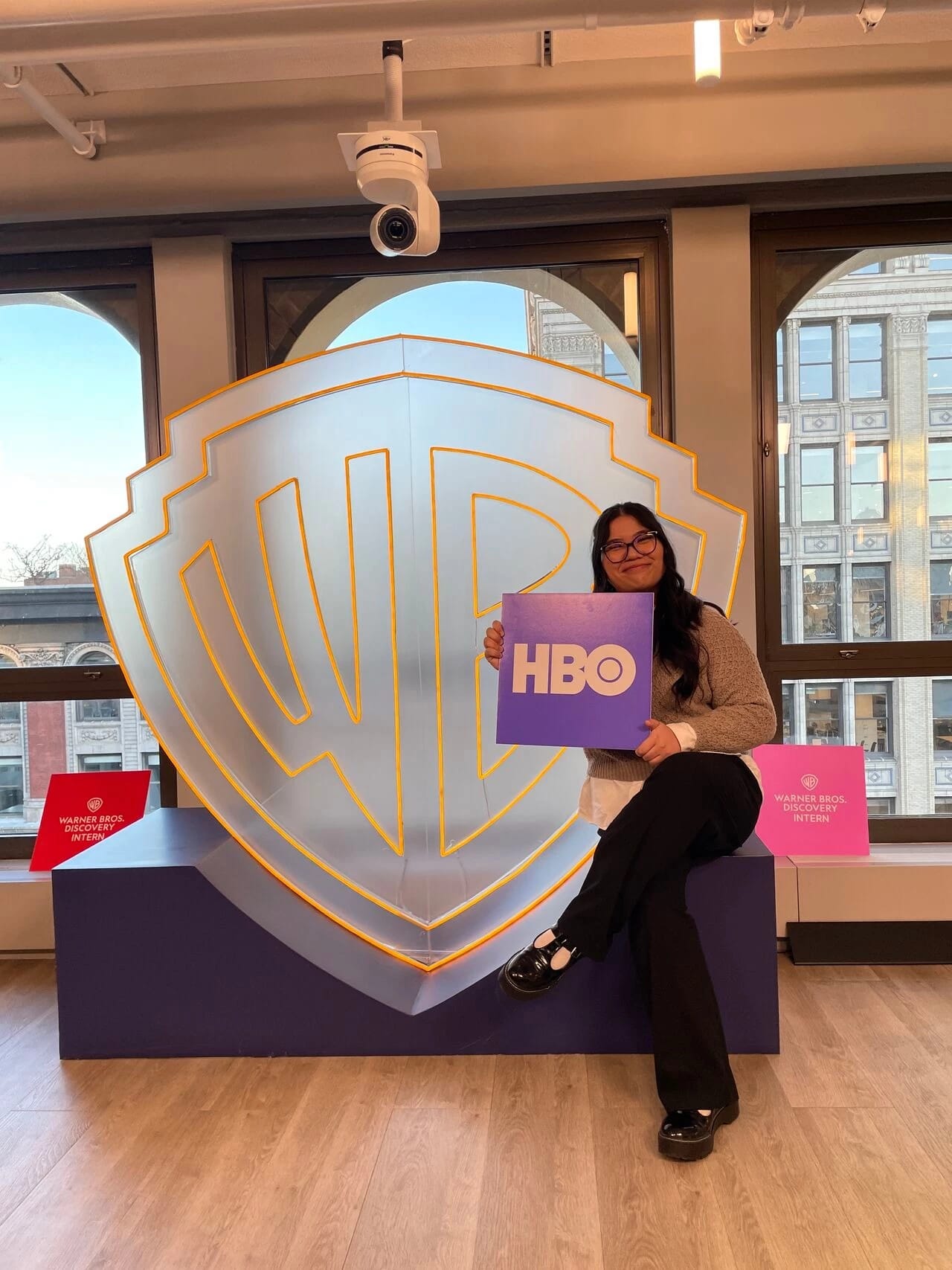 Student holding HBO logo sign in front of larger transparent Warner Brothers logo sign.