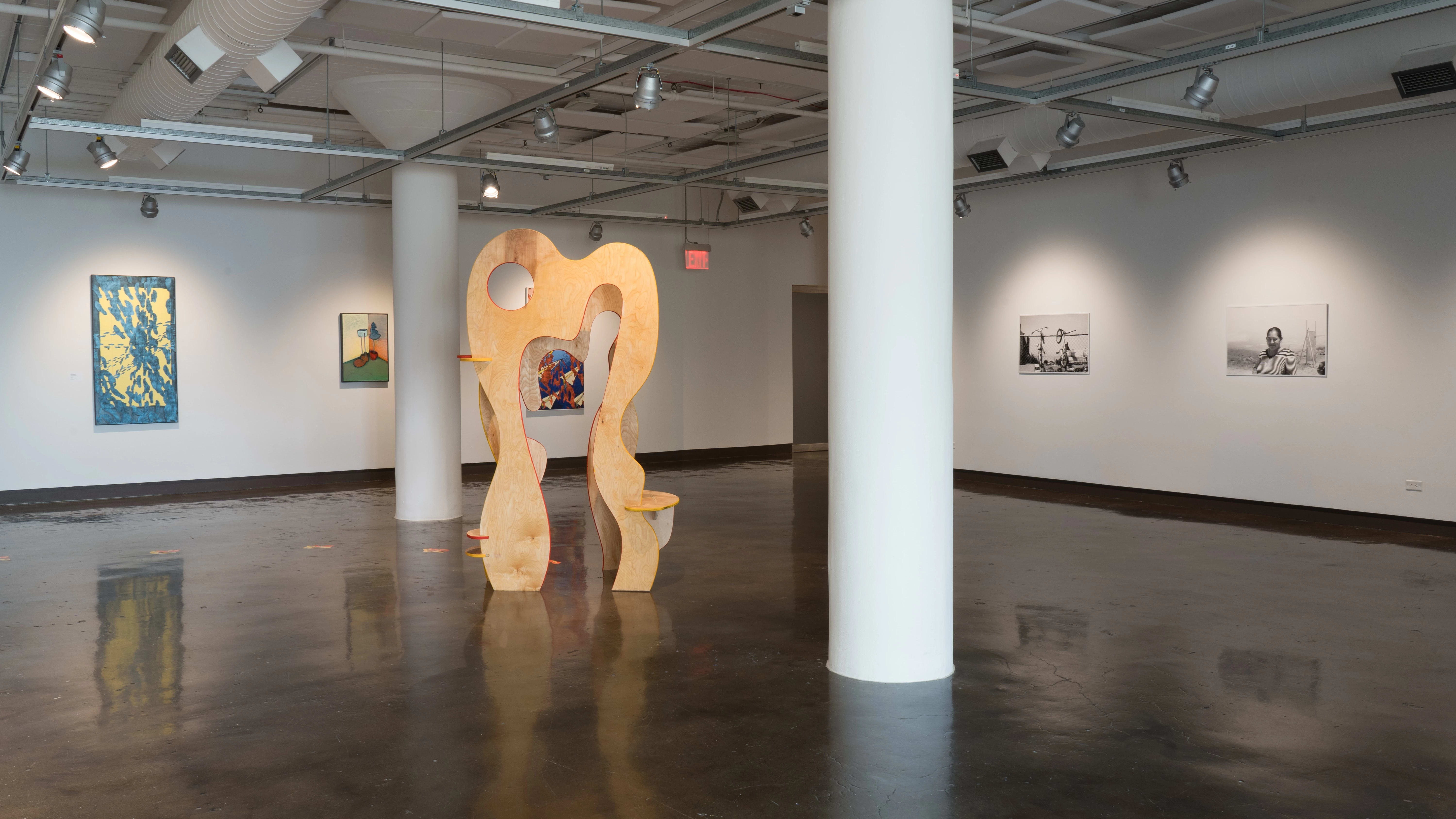 In the first gallery space, a large wood sculpture stands in the middle of the room. Surrounding this sculpture are black-and-white inkjet prints and oil paintings.
