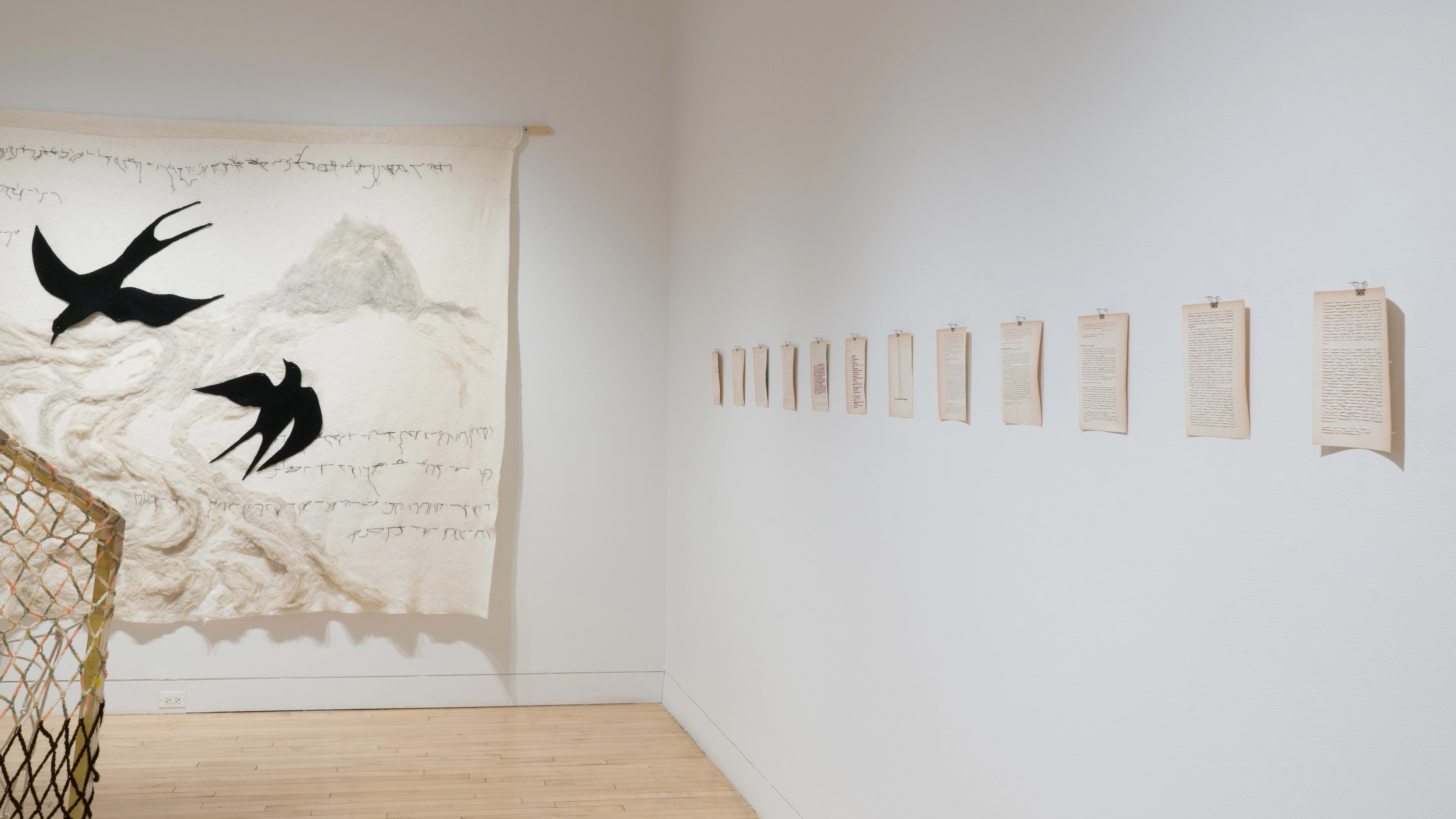 Another view of the west and south walls in the Flatiron Gallery, showing a wooden house-like structure with rope walls on the floor. Papers from a book are pinned to the wall on the right, and a large white tapestry with black swallows hangs on the south wall.