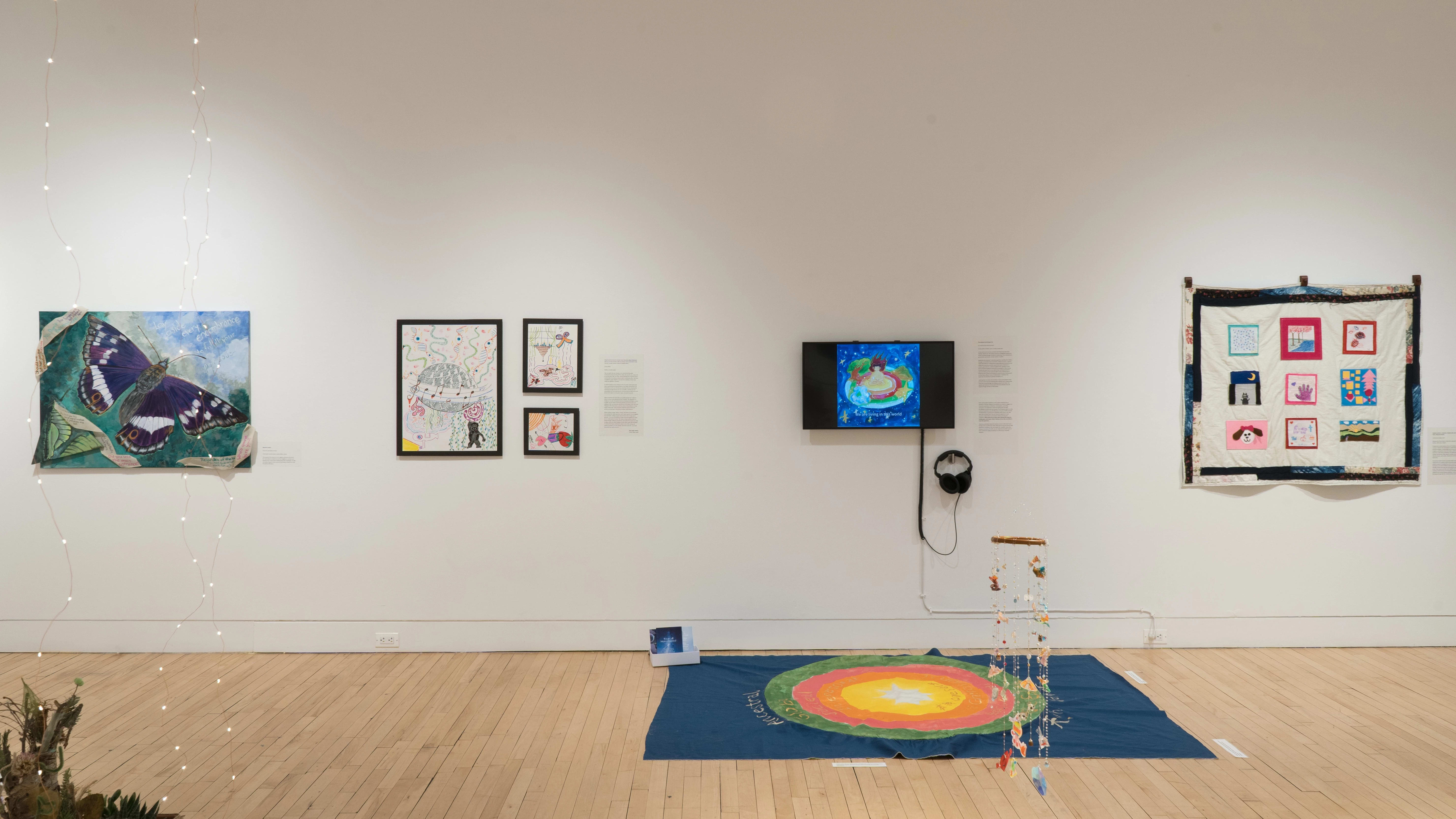 Straight shot of the east wall of the Flatiron Gallery. We see two large tapestry pieces, one on the wall and one on the floor in the foreground; a monitor on the middle, and a selection of drawings to the left, followed by a large butterfly painting.