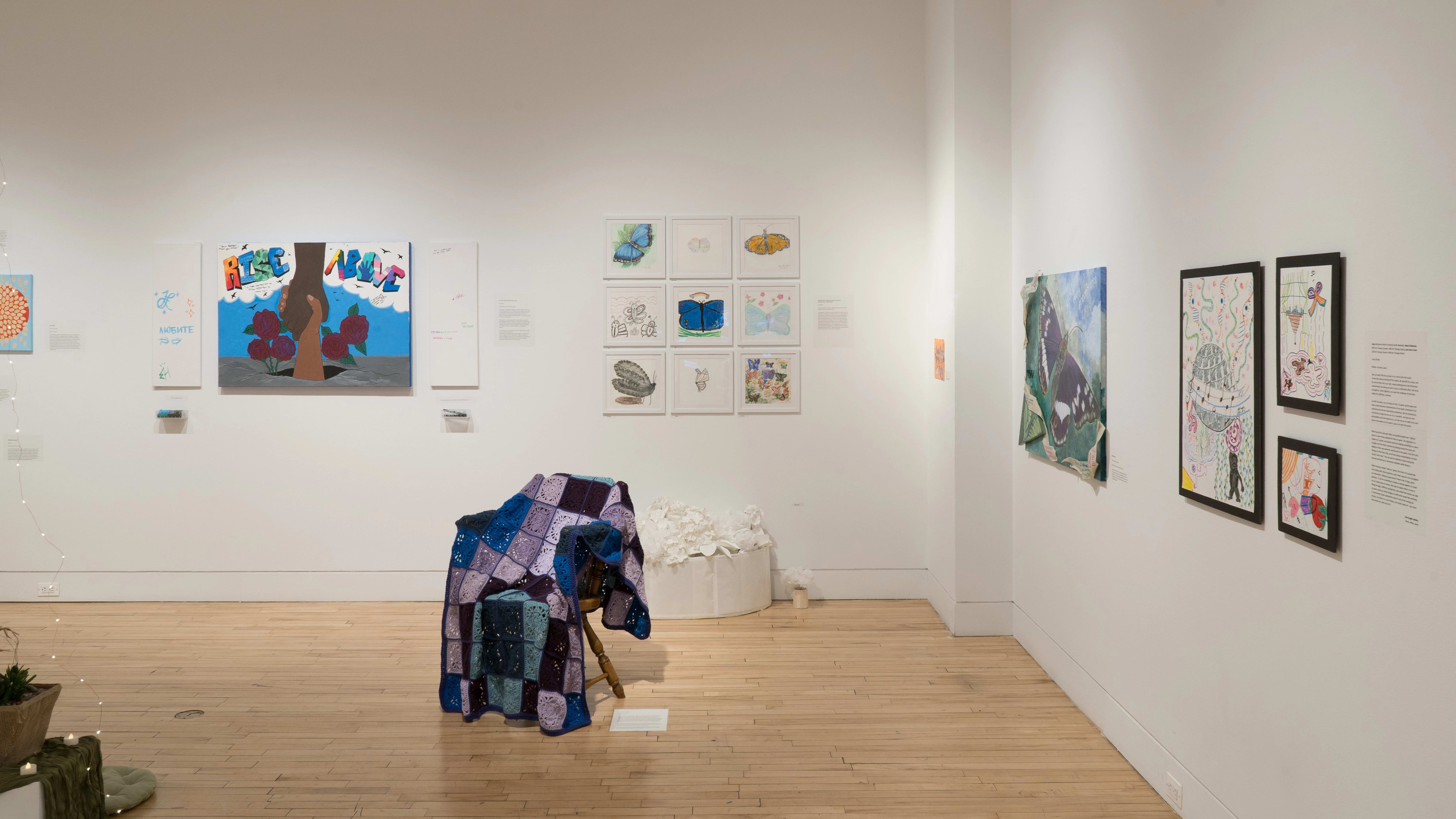 Straight shot of the north and east walls of the Flatiron Gallery. We see a selection of drawings to the right, followed by a large butterfly painting on the east wall. On the north wall, we see a grid of colorful drawings on the right and a chair with a crochet blanket draped over it in the foreground.