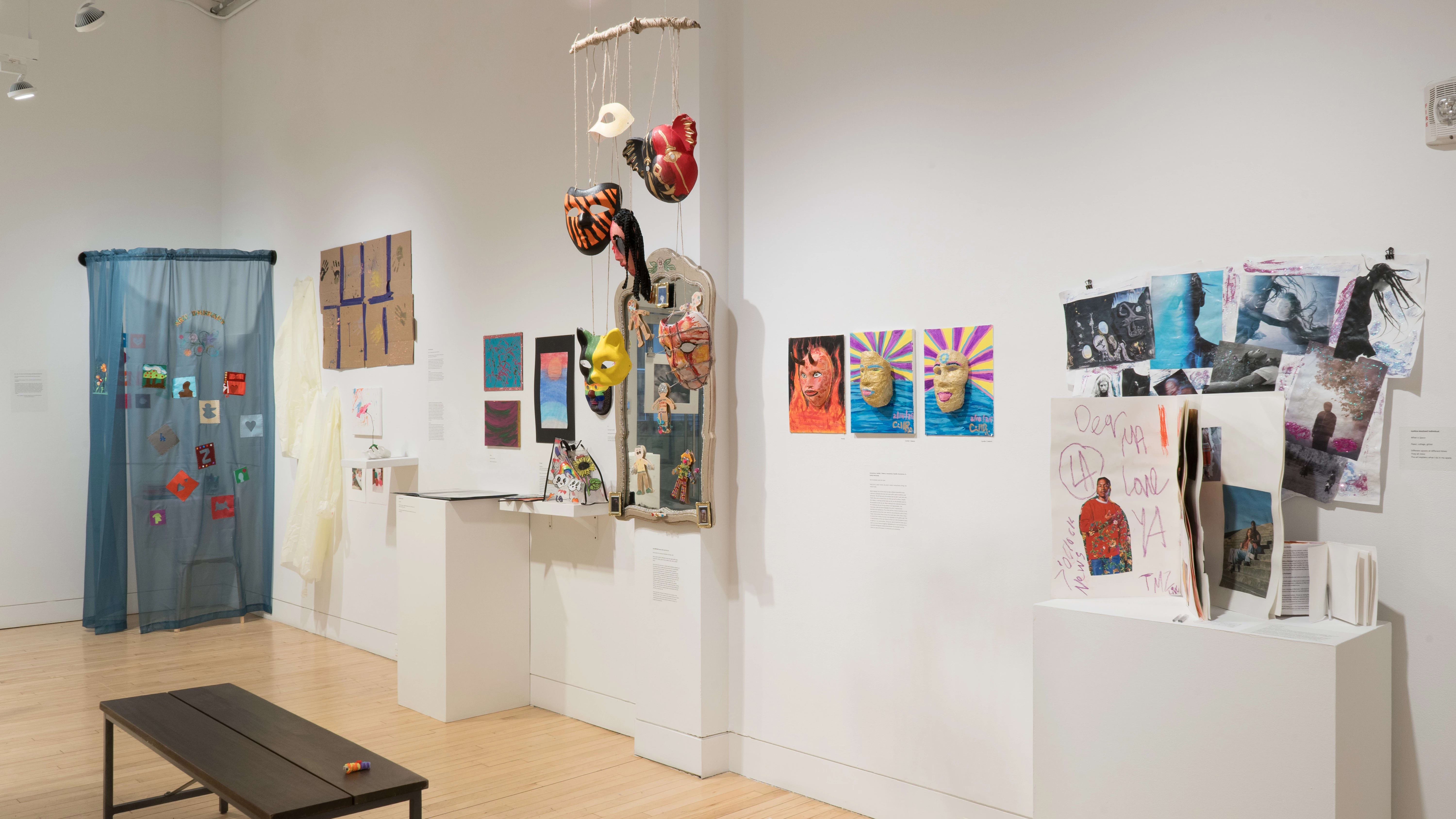 Angled shot of the west wall. We see many mixed media installations, with a mobile of masks hanging from the ceiling and a pedestal of paper works in the foreground. There is a large blue curtain installation in the back corner.