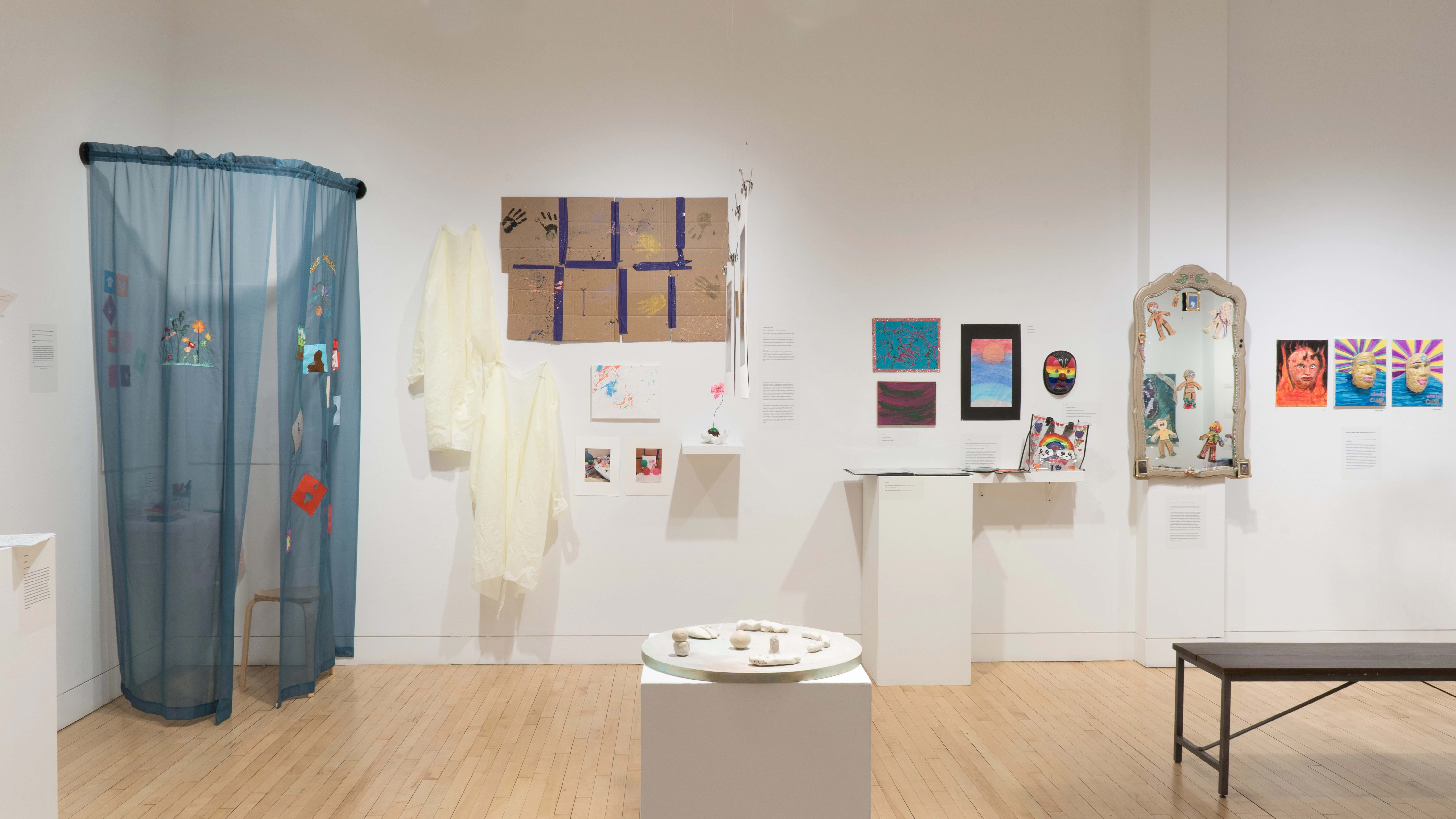 Straight shot of the west wall. We see many mixed media installations, with a pedestal of small ceramics in the foreground. There is a large blue curtain installation in the back corner, and a large mirror on the column.