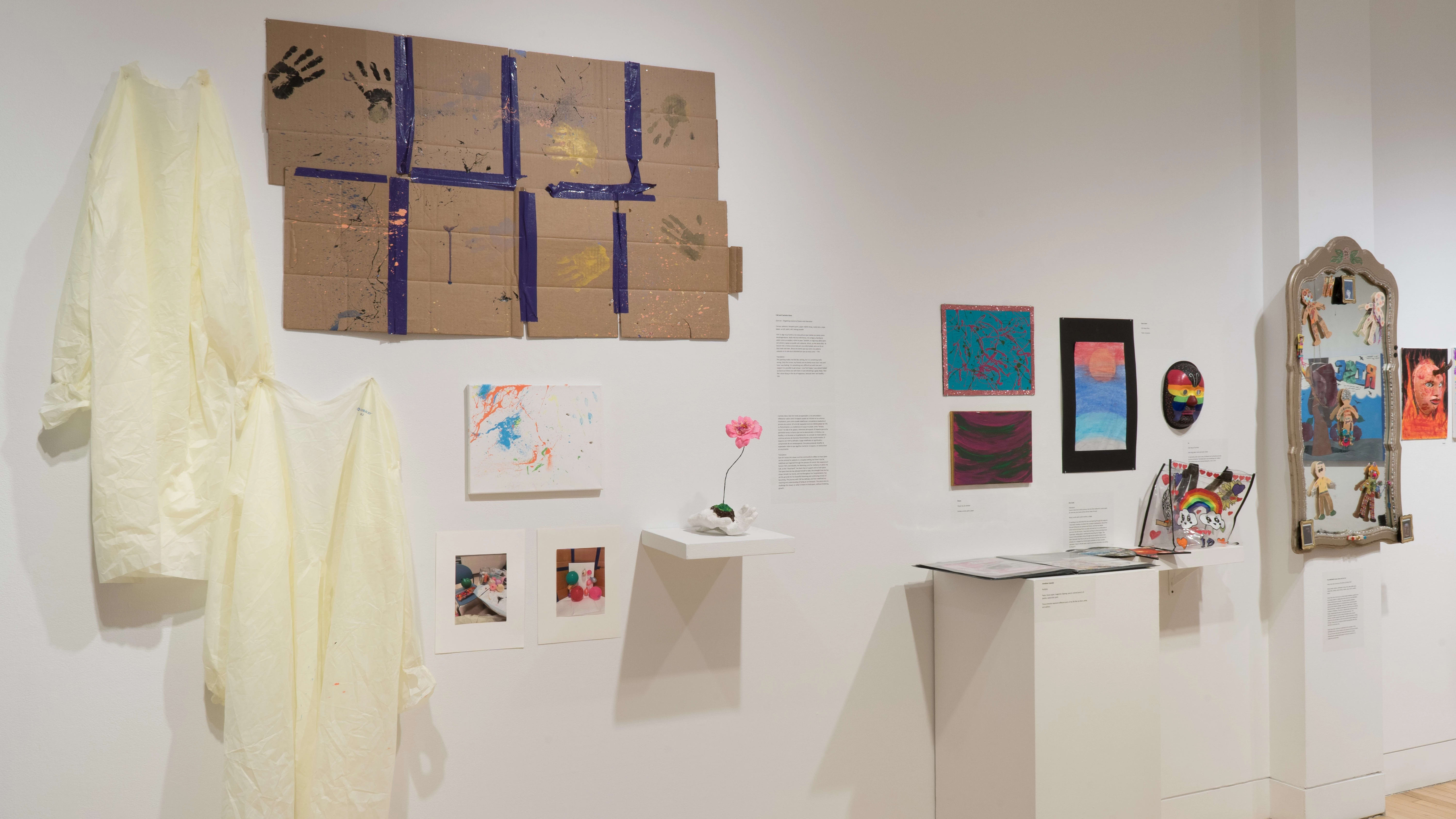 Angled shot of the west wall. There are two hanging garments in the foreground, a pedestal against the wall, and paper works on the walls. There is a large mirror on the column in the background.