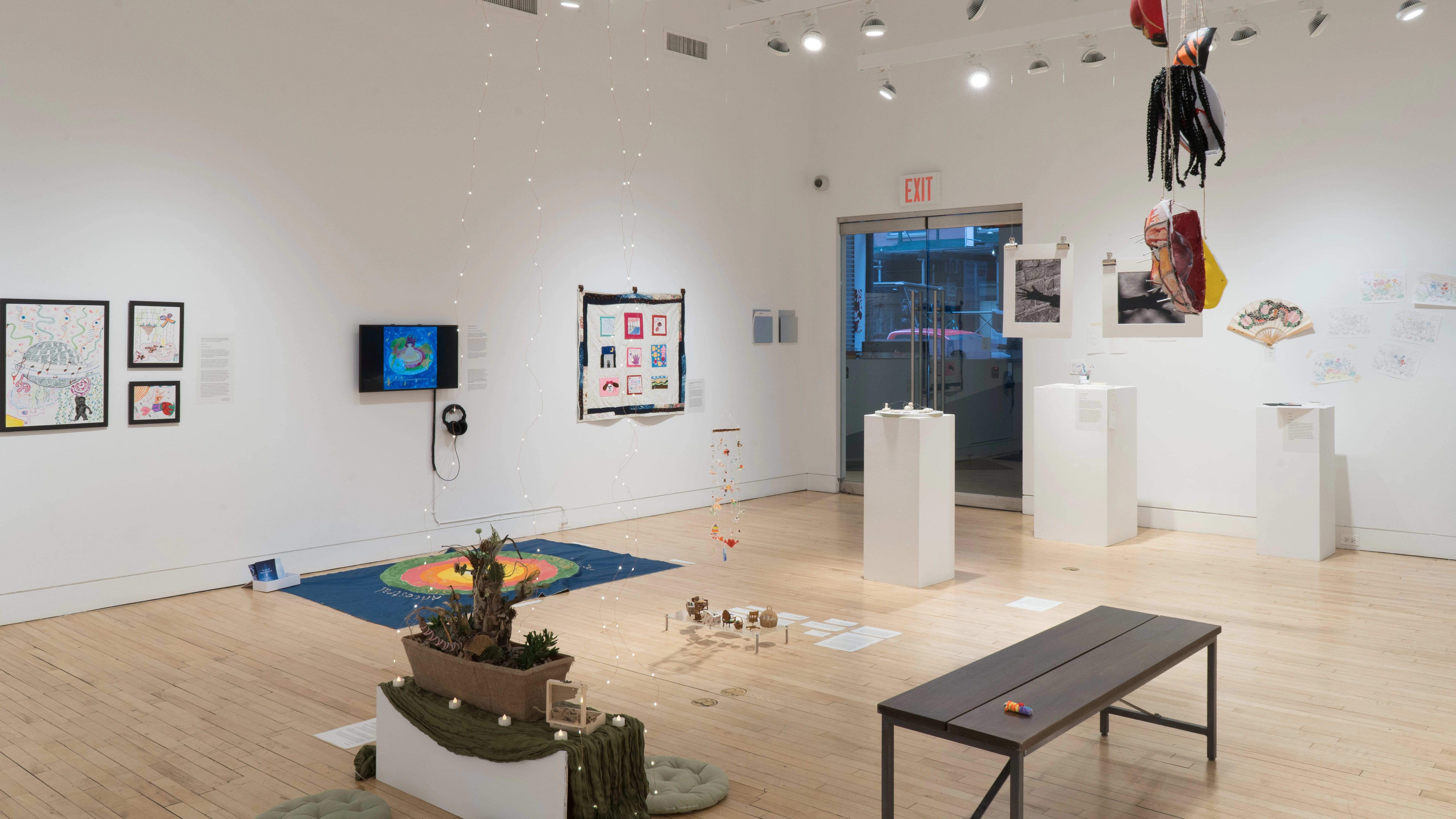 Overview of the Flatiron Gallery facing southeast. There are many works of varying sizes spread throughout, lining the walls, hanging from the ceiling, and scattered on the floor. The gallery doors are visible in the background.