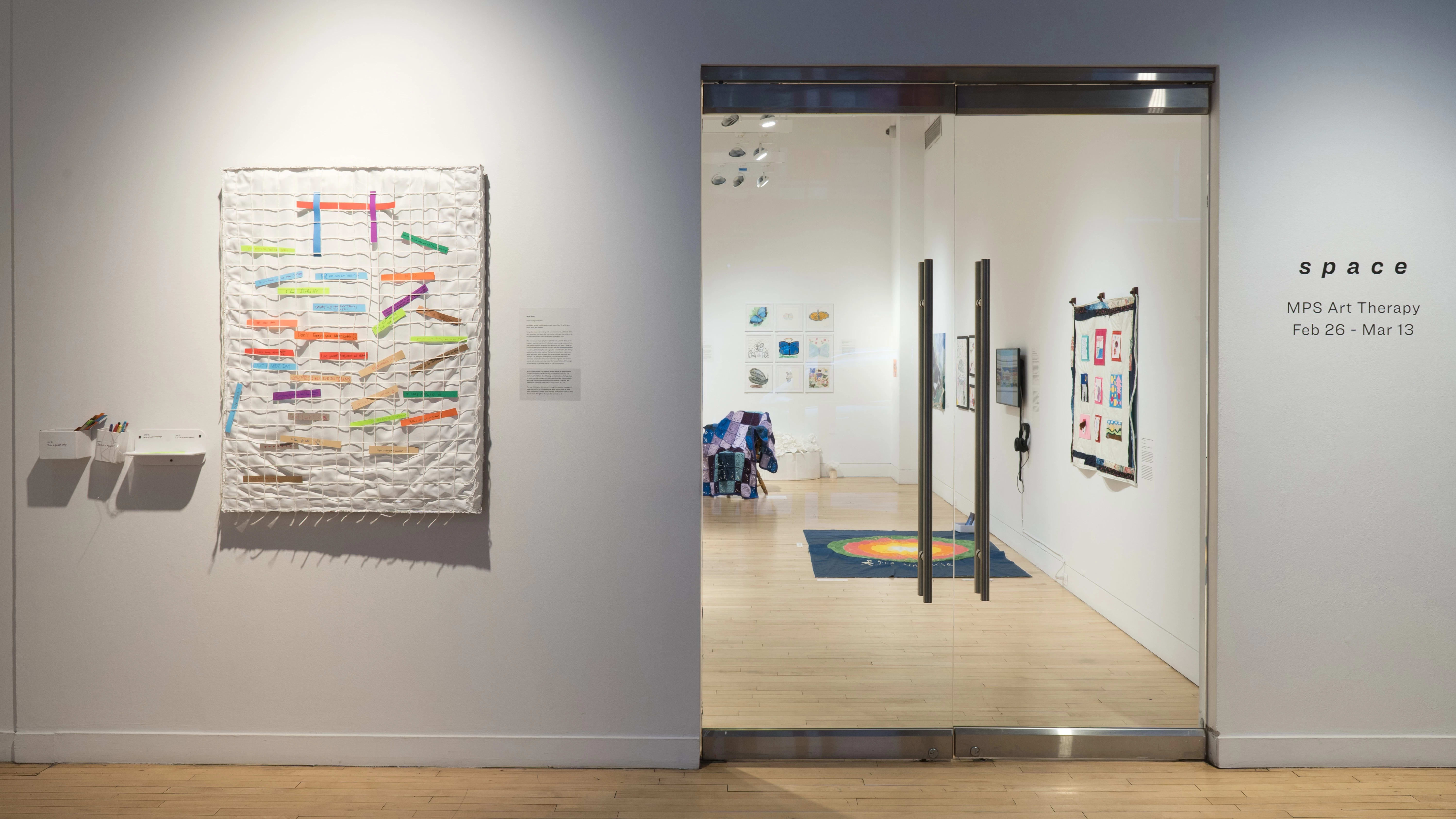 View of the alcove at the Flatiron Gallery, The glass gallery doors are visible with “space” written in vinyl to the right. There’s a large textile piece on the wall to the left of the doors.