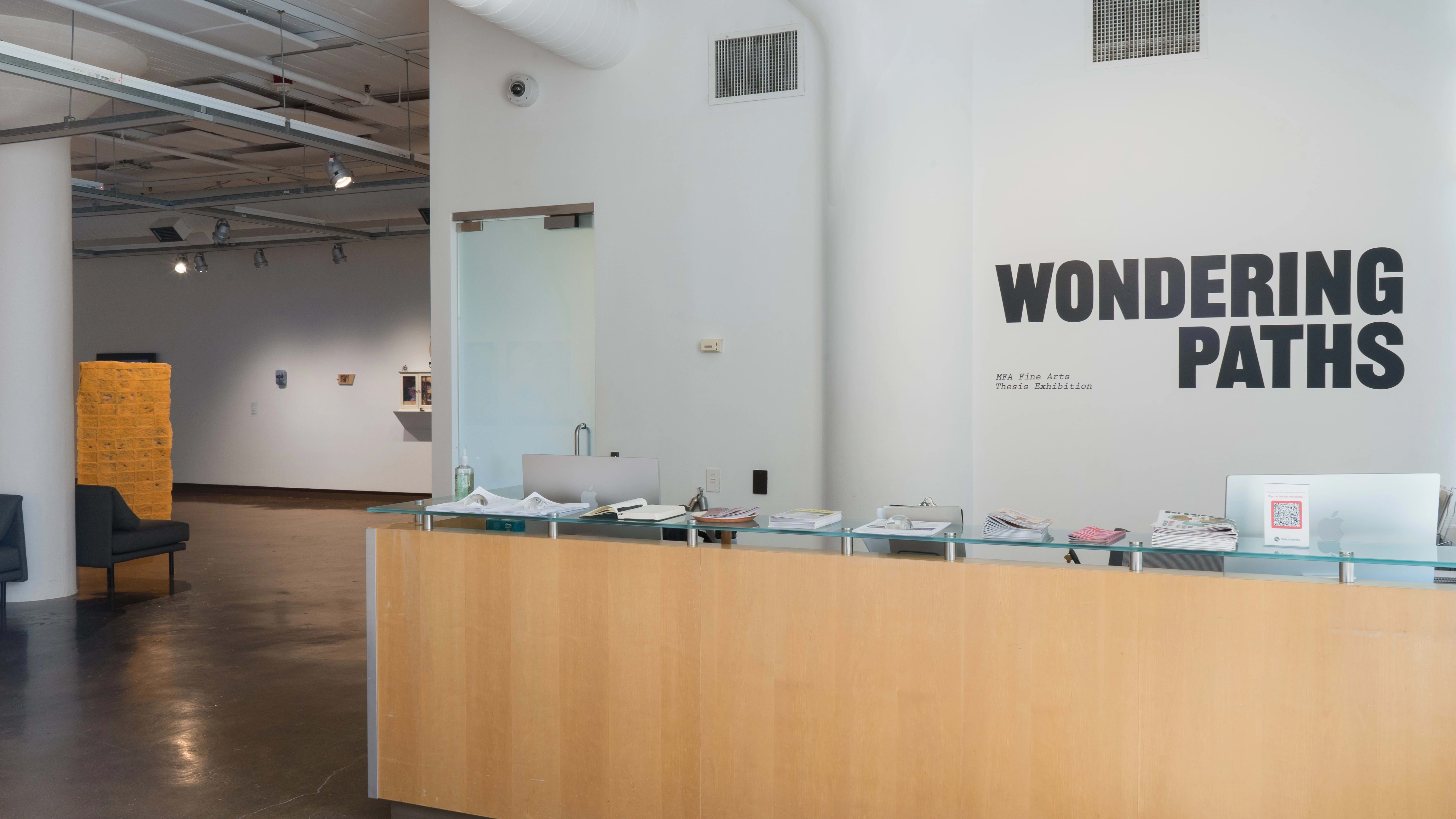 “Wondering Paths,” the title of the 2025 MFA Fine Arts thesis exhibition, is displayed in wall vinyl over the front desk of the SVA Chelsea Gallery. Works are visible in the gallery beyond the desk.