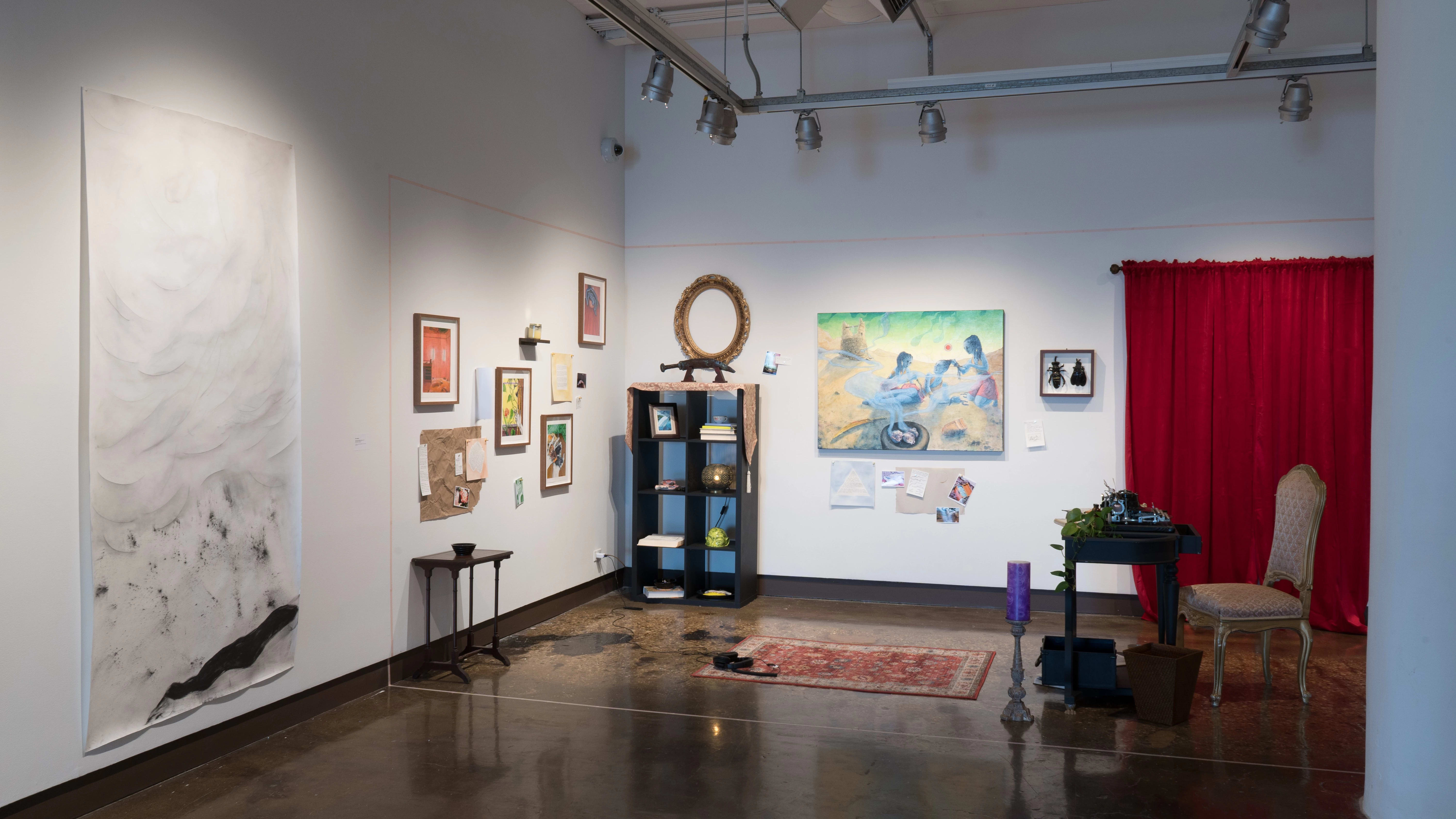 A room-like installation is set up in the corner of the space, with a large painting, a desk, a candle, a bookshelf, and other objects. To the left of the installation, a charcoal drawing on a long, vertical, mostly white piece of paper is hung on the wall.