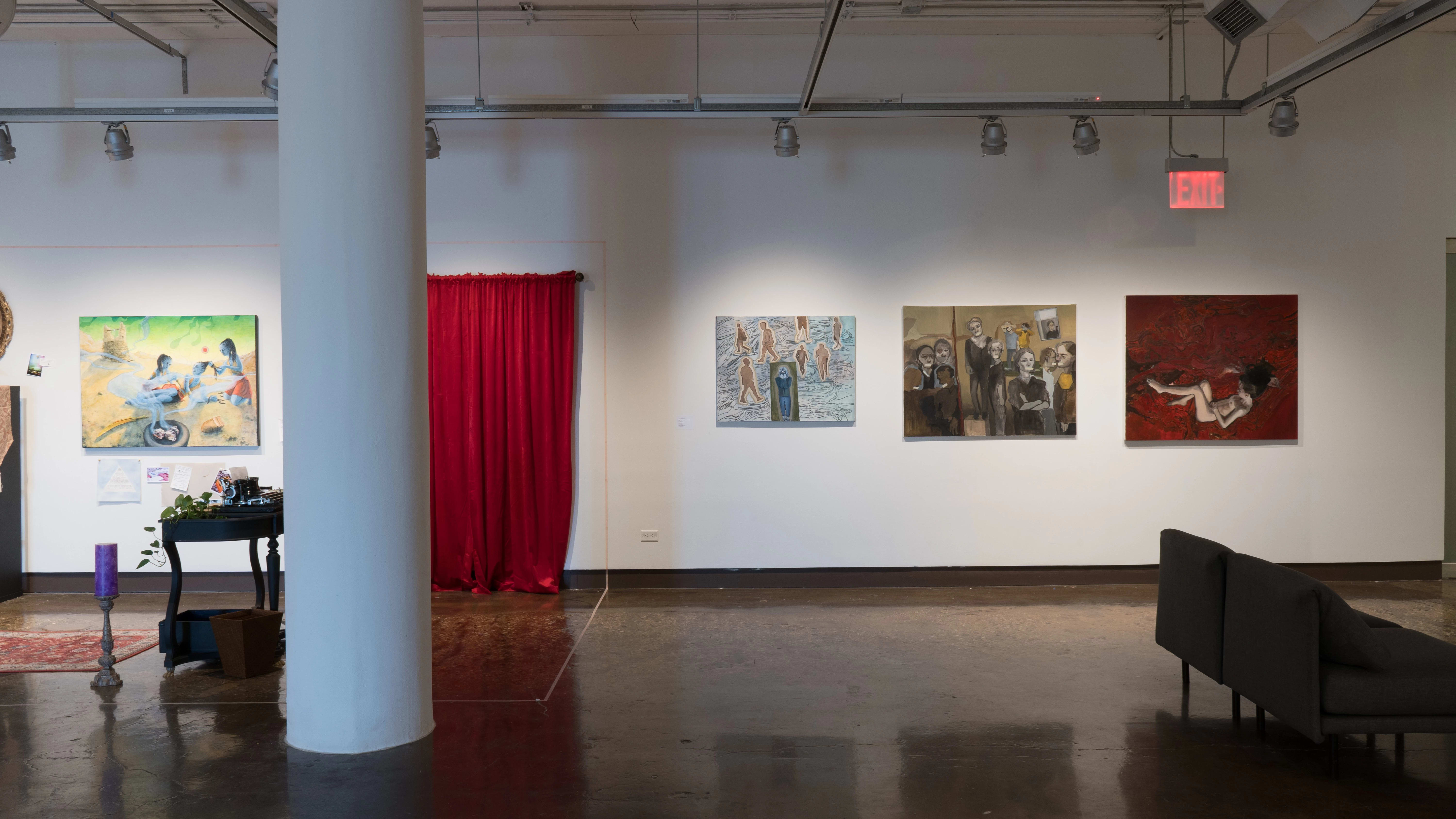 On the left, a room-like installation is set up in the corner of the space, with a large painting, a desk, a candle, and a red velvet curtain. To the right of this installation, three paintings on canvas are hung in a row.