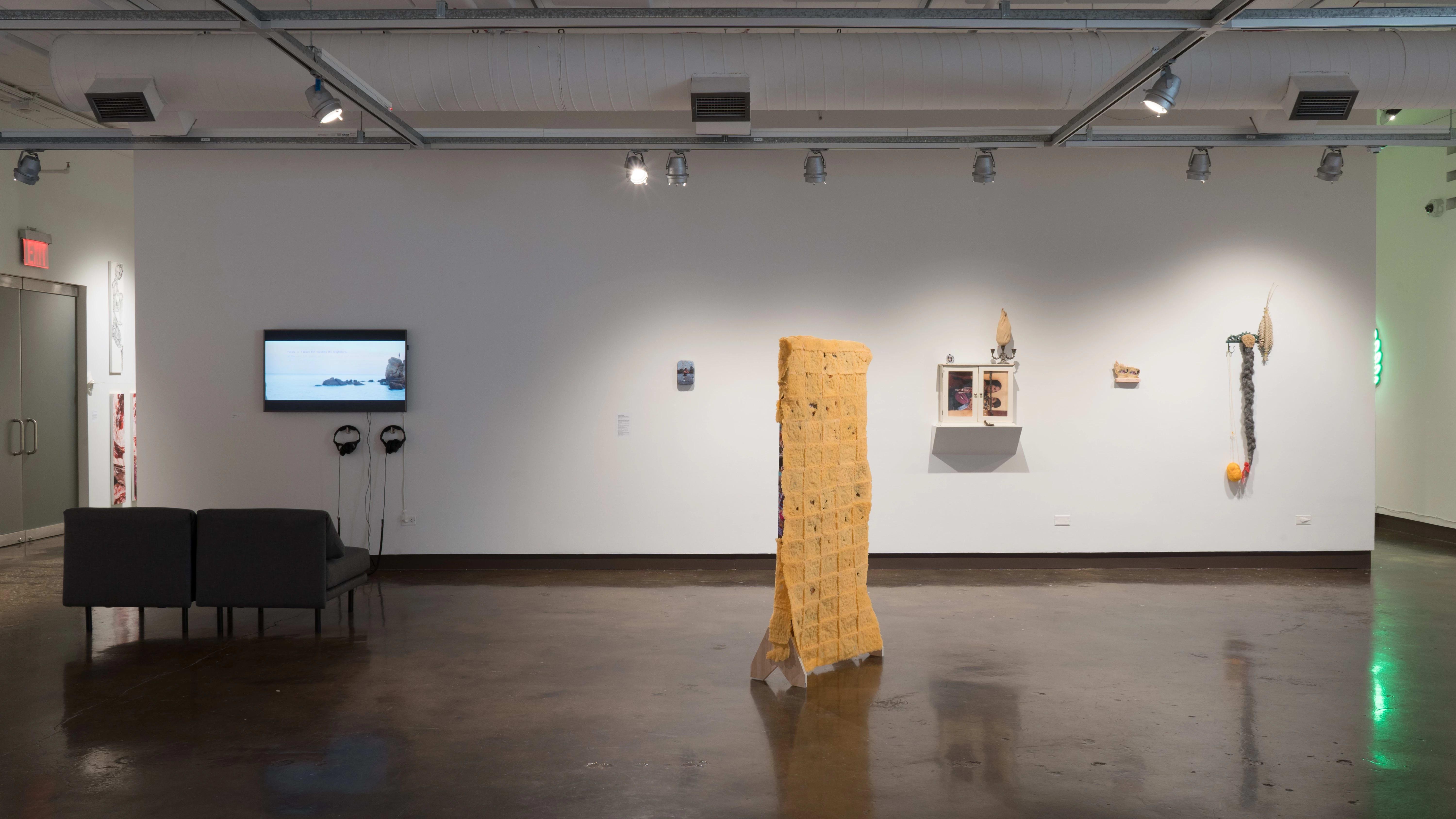A wooden structure coated in spongy material stands in the center of the room. To its left, two chairs sit in front of a monitor. To its right, assemblages using cabinets, photos, candlesticks, a long braid, and other objects are mounted on the wall.