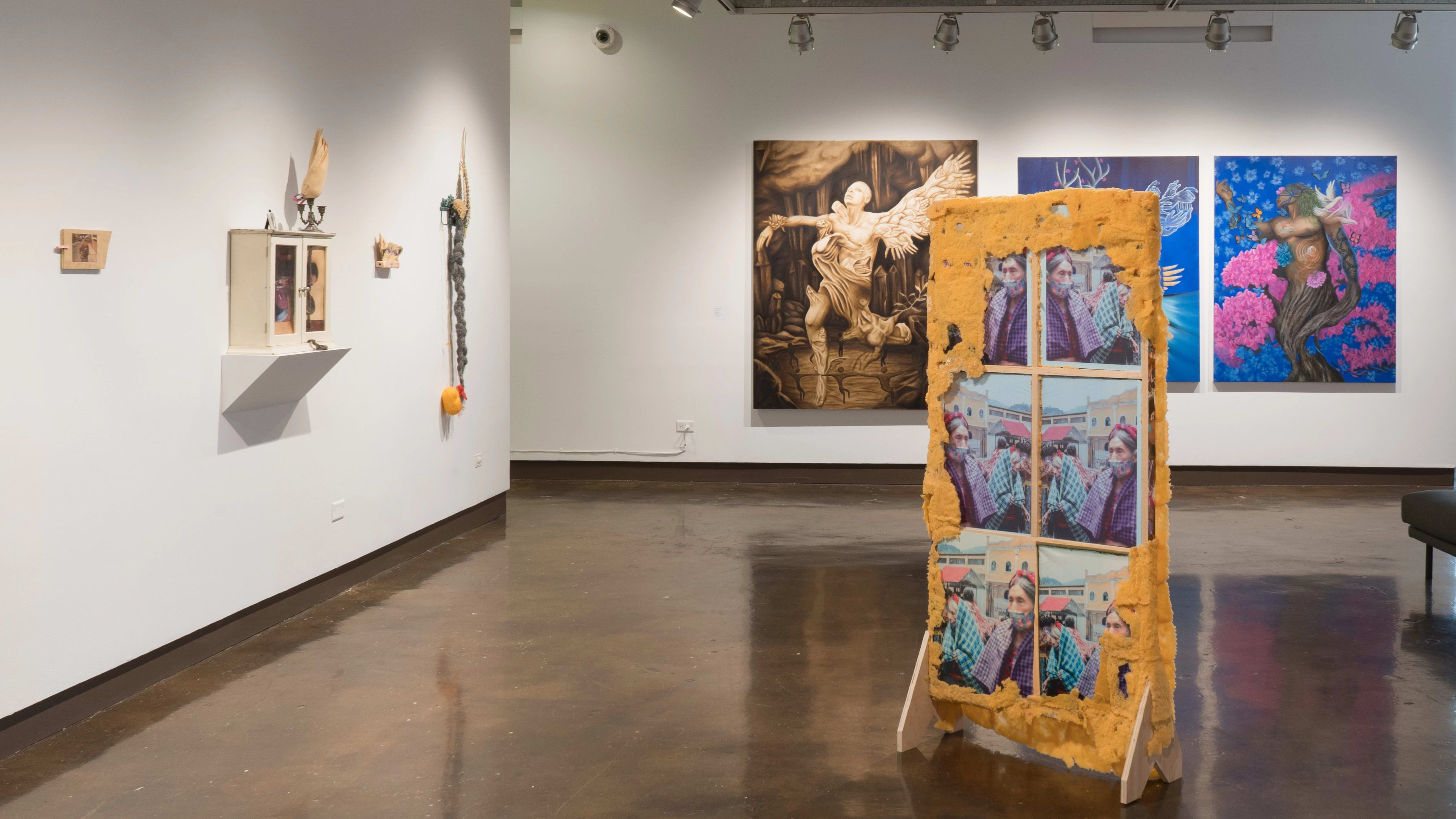 A wooden structure standing on the floor of the gallery is coated in an orange, spongy material and frames a photo of an older woman. The large figurative paintings can be seen on the wall behind it, while mixed media sculptures and assemblages hang on the wall to the left.
