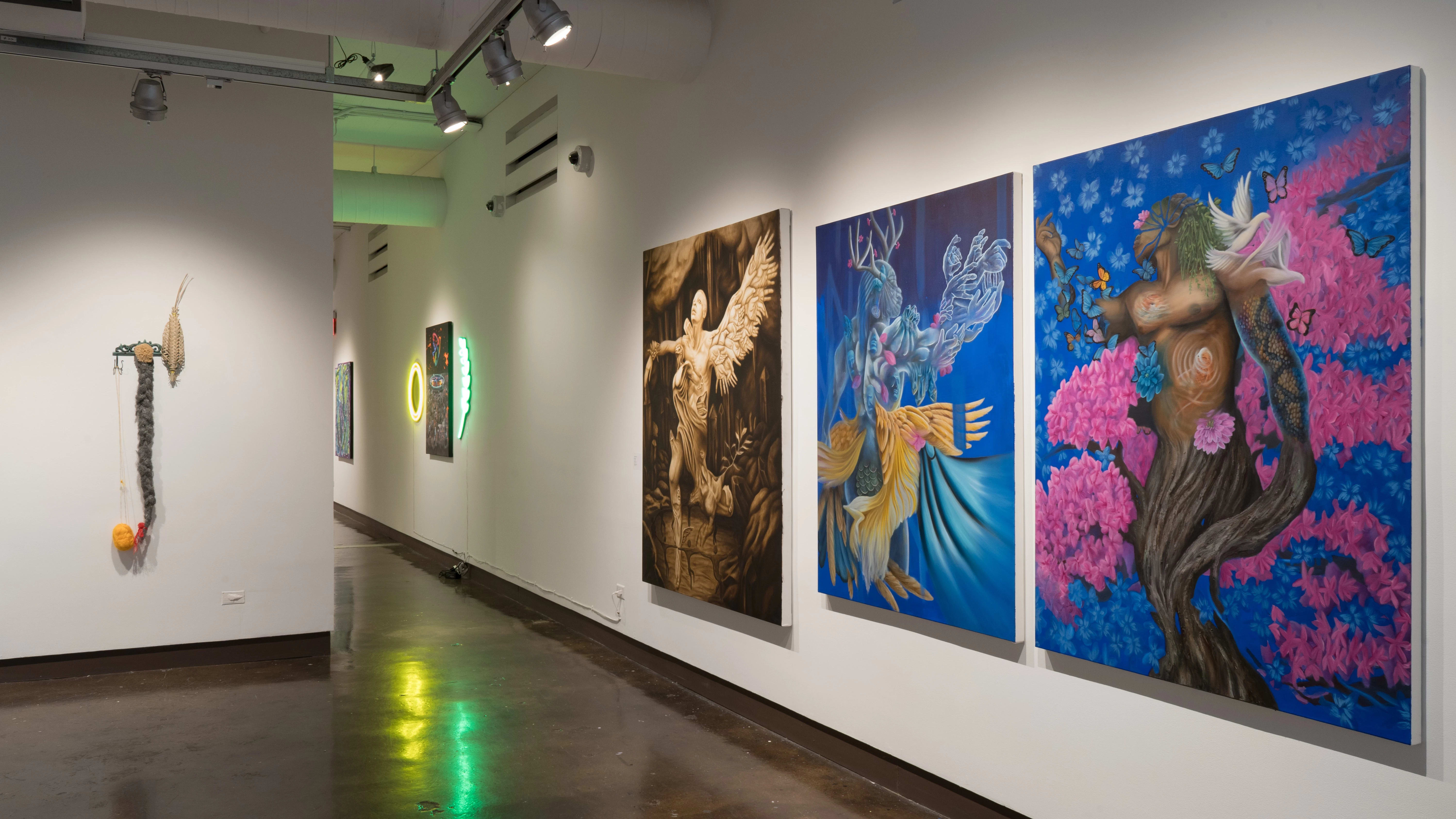 On the right wall of gallery one, three large paintings depict human figures merging with nature in various states. One painting is monotone sepia, while the other two are a vibrant cobalt with pinks and yellows. A mixed media assemblage is visible on the wall to the left.