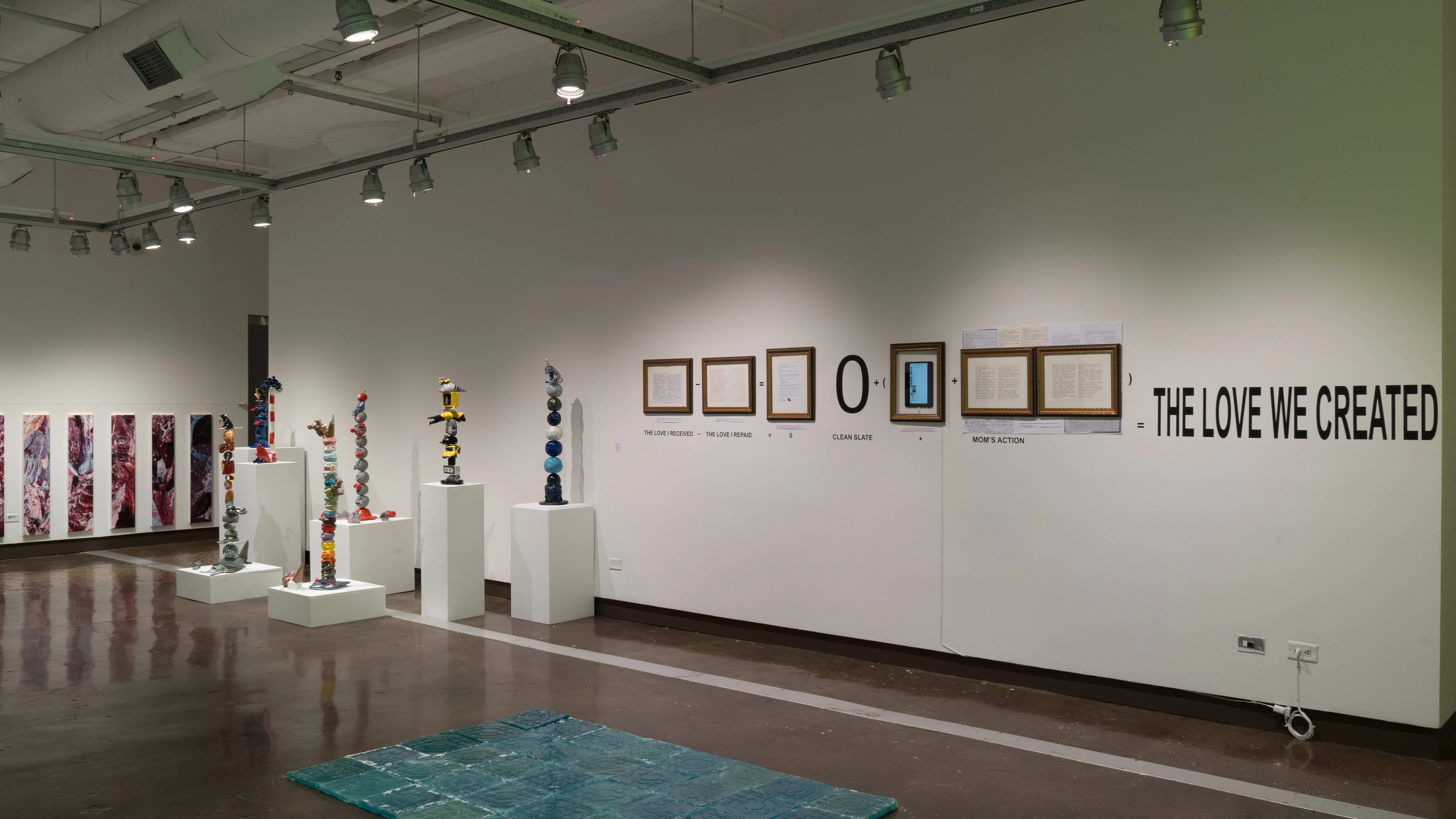 In gallery two, the meat paintings, the precarious ceramic sculptures, and the blue tiles are all visible around a row of frames on the wall. With the frames, there is text, showing the frames as parts of an equation, which equals out to the phrase “the love we created.”