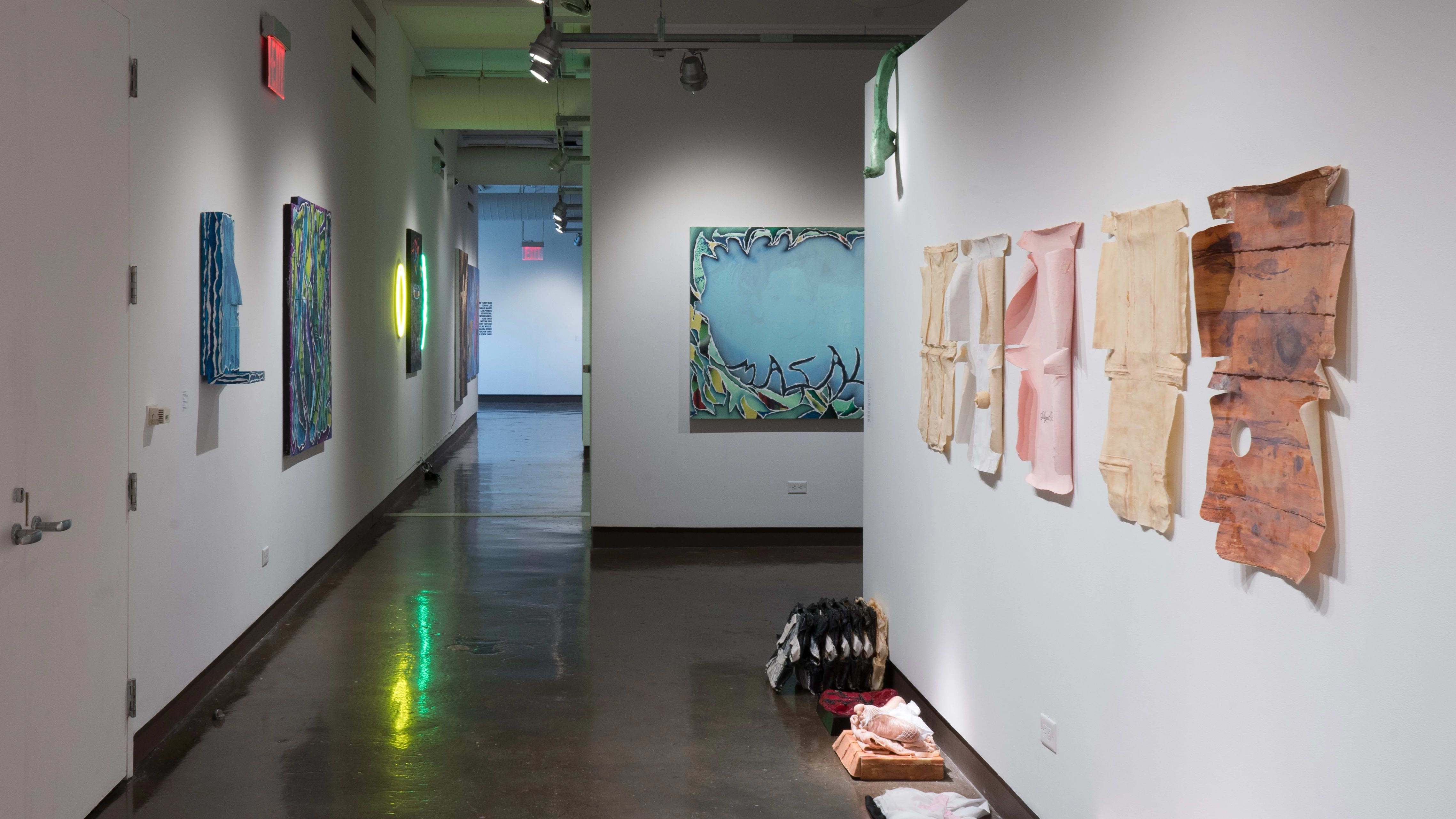 Looking back toward gallery one through the right hallway, some paper works displayed to the immediate right are seemingly peeling off the wall. Sculptures sit on the ground beneath them, while large paintings are visible on the left wall and in gallery 3 beyond them.
