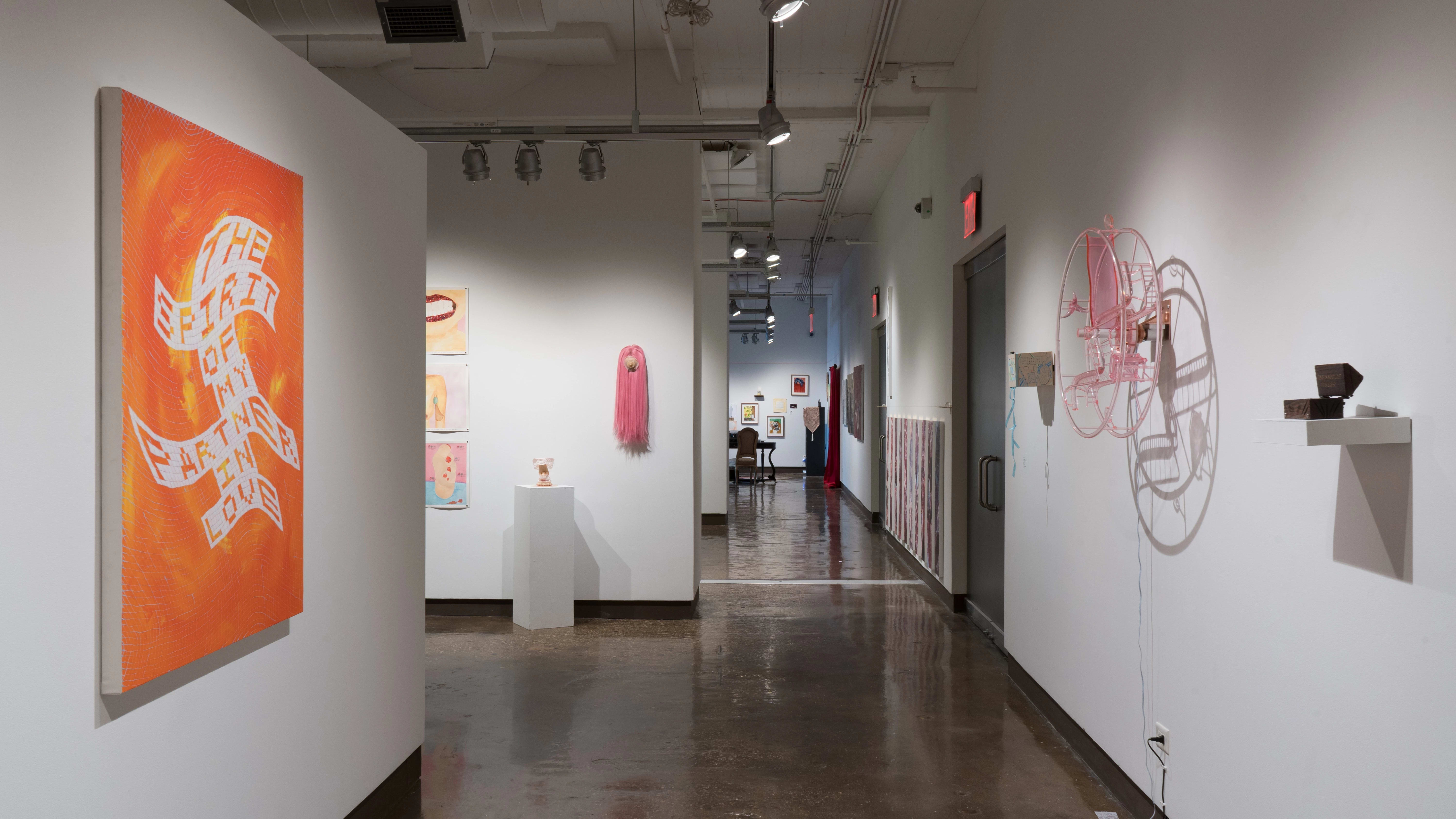 Looking back toward gallery one through the left hallway, an orange painting featuring blocky text is hung to the immediate left. To the right, a pink acrylic wheel is mounted on the wall beside a small wooden box. More works are visible in galleries three, two, and one.