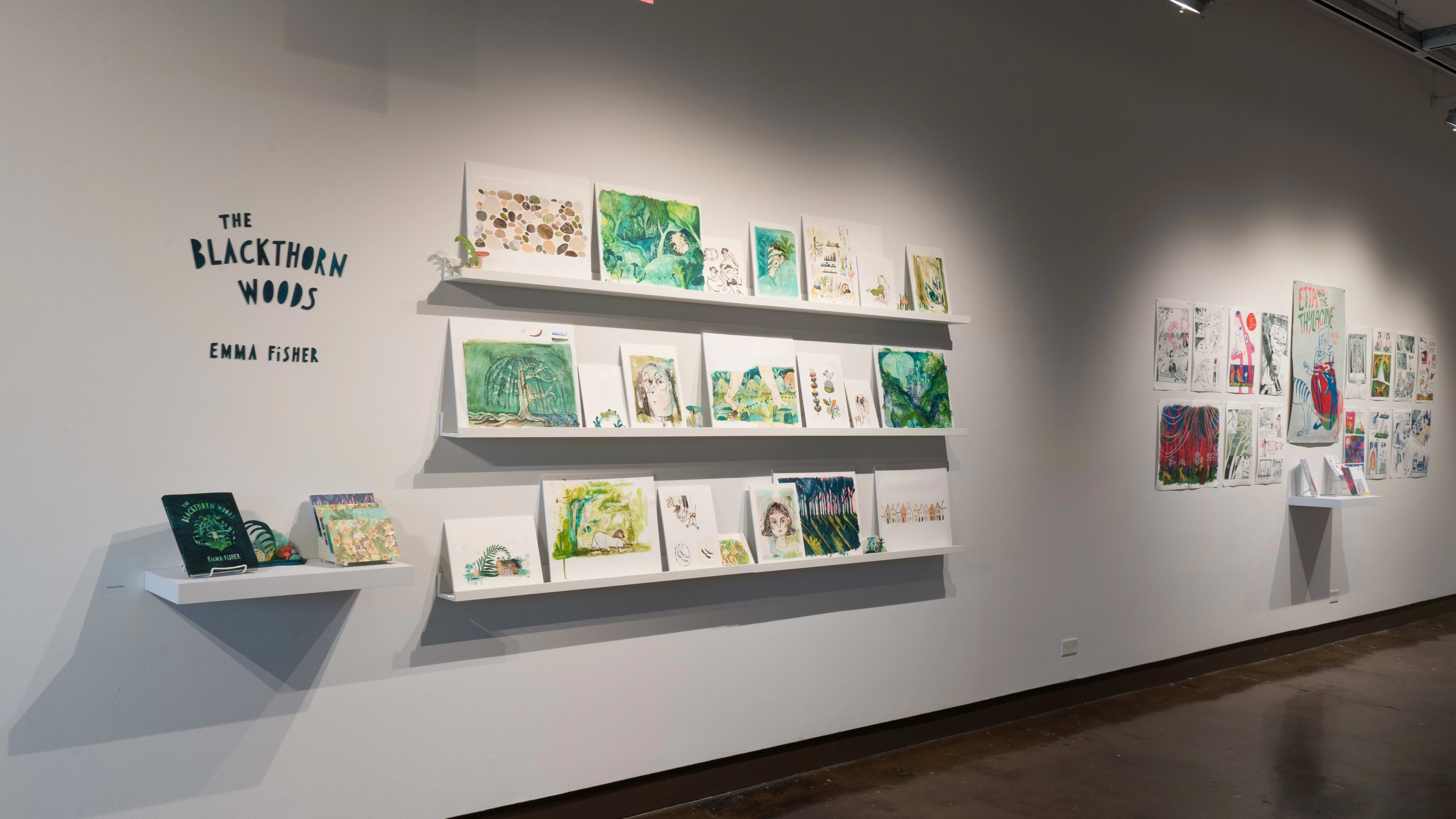 Angled view of the west wall at the SVA Chelsea Gallery. Three shelves of watercolor paintings in blues and greens hang in the foreground, and a small shelf with books and cards hangs below the title, “The Blackthorn Woods, Emma Fisher.”